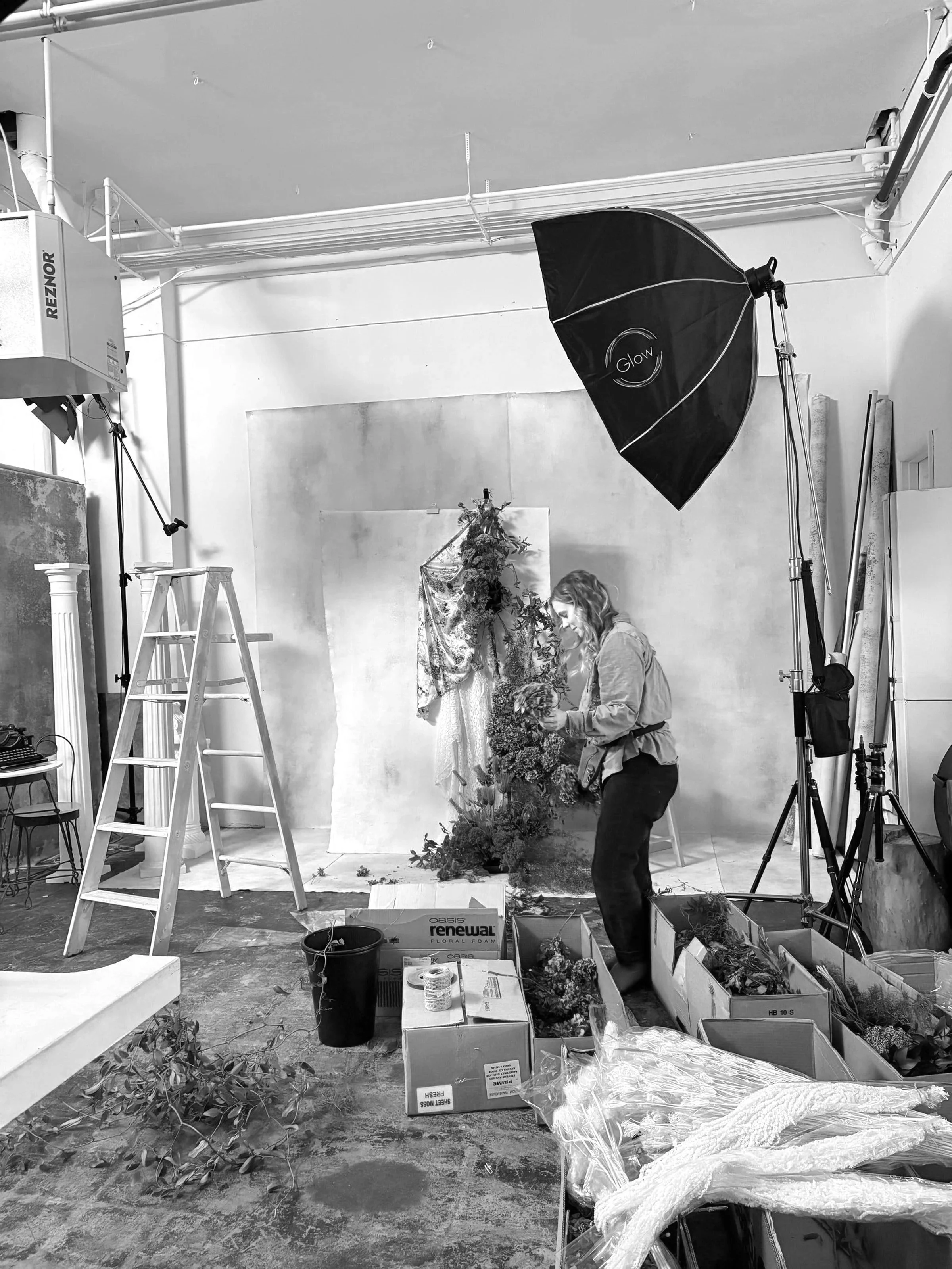 Black and white photo of a woman working on a floral arrangement in a studio. The studio has studio lighting equipment, a ladder, and cardboard boxes with flowers and plant materials.