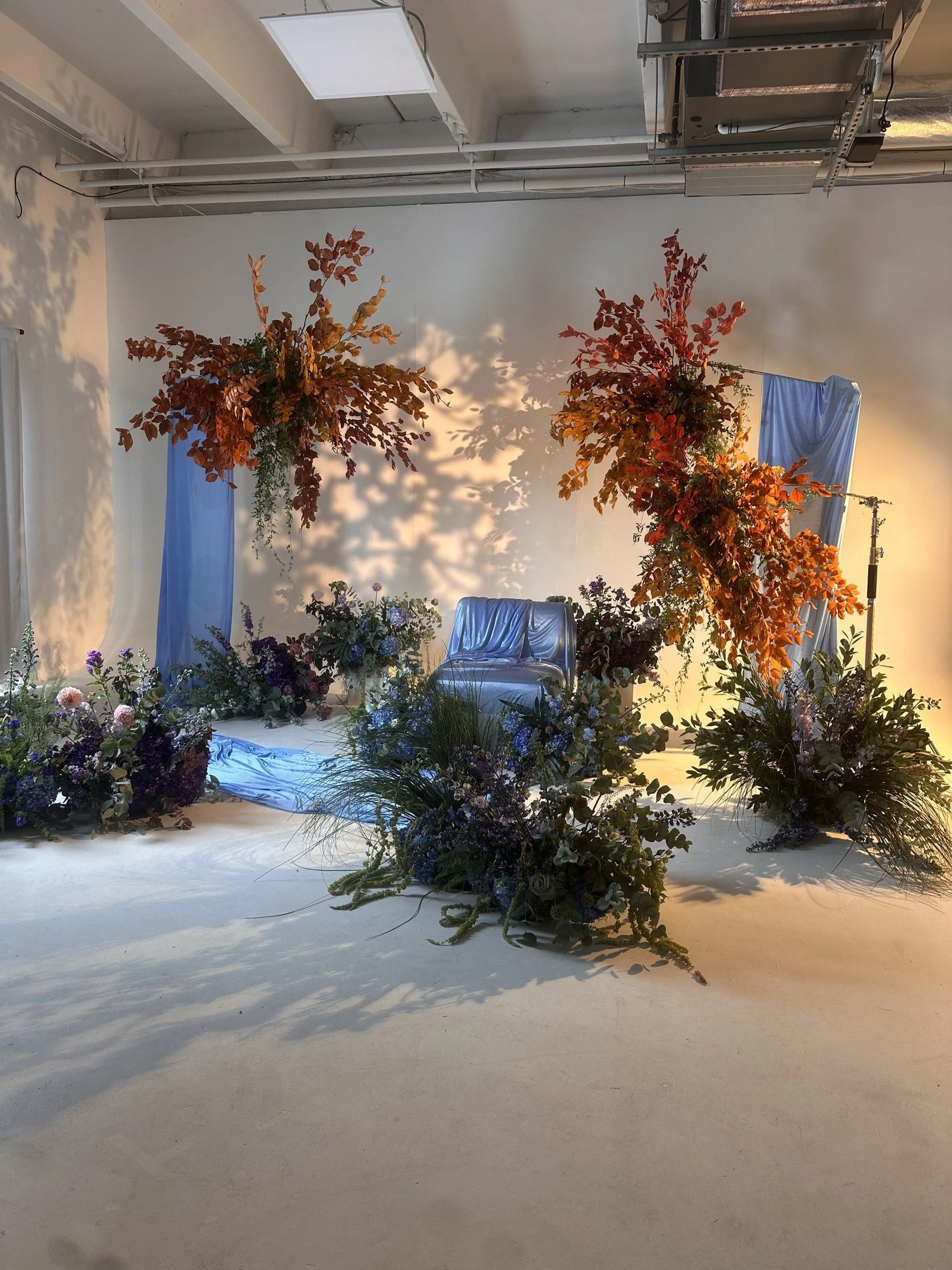 A decorated indoor space with floral arrangements, orange and purple flowers, blue fabric drapery, and a blue chair, likely set up for an event or photoshoot.