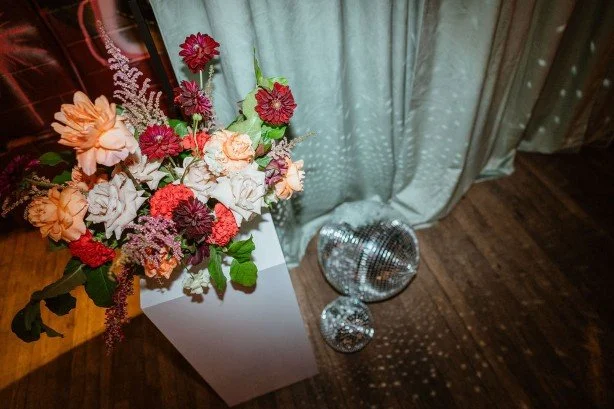 Colorful flower arrangement in a white vase, positioned on a floor next to two disco balls, in front of a green curtain.