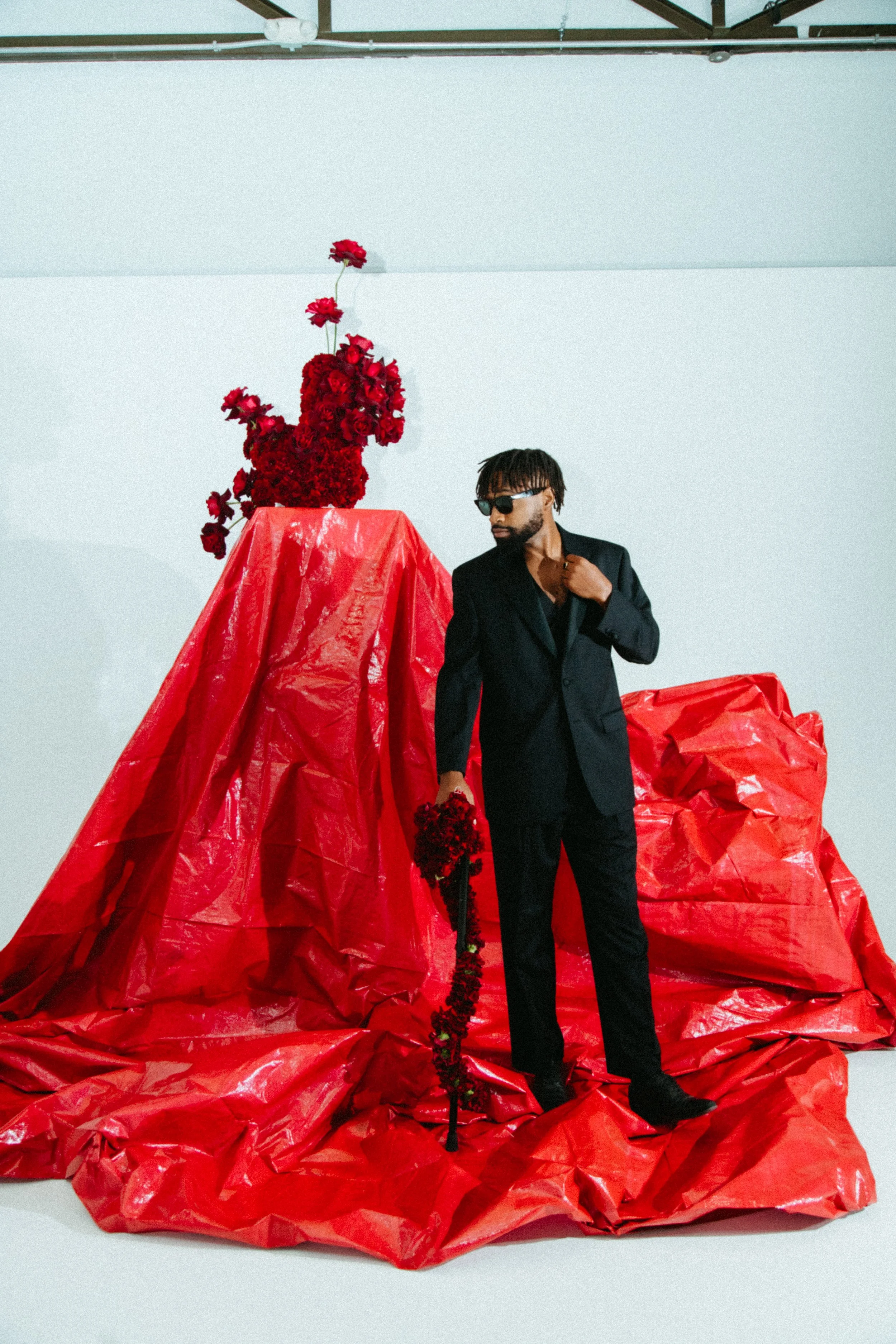 a Black male model in a dark suit against a background of white with a red backdrop and red carnations and roses