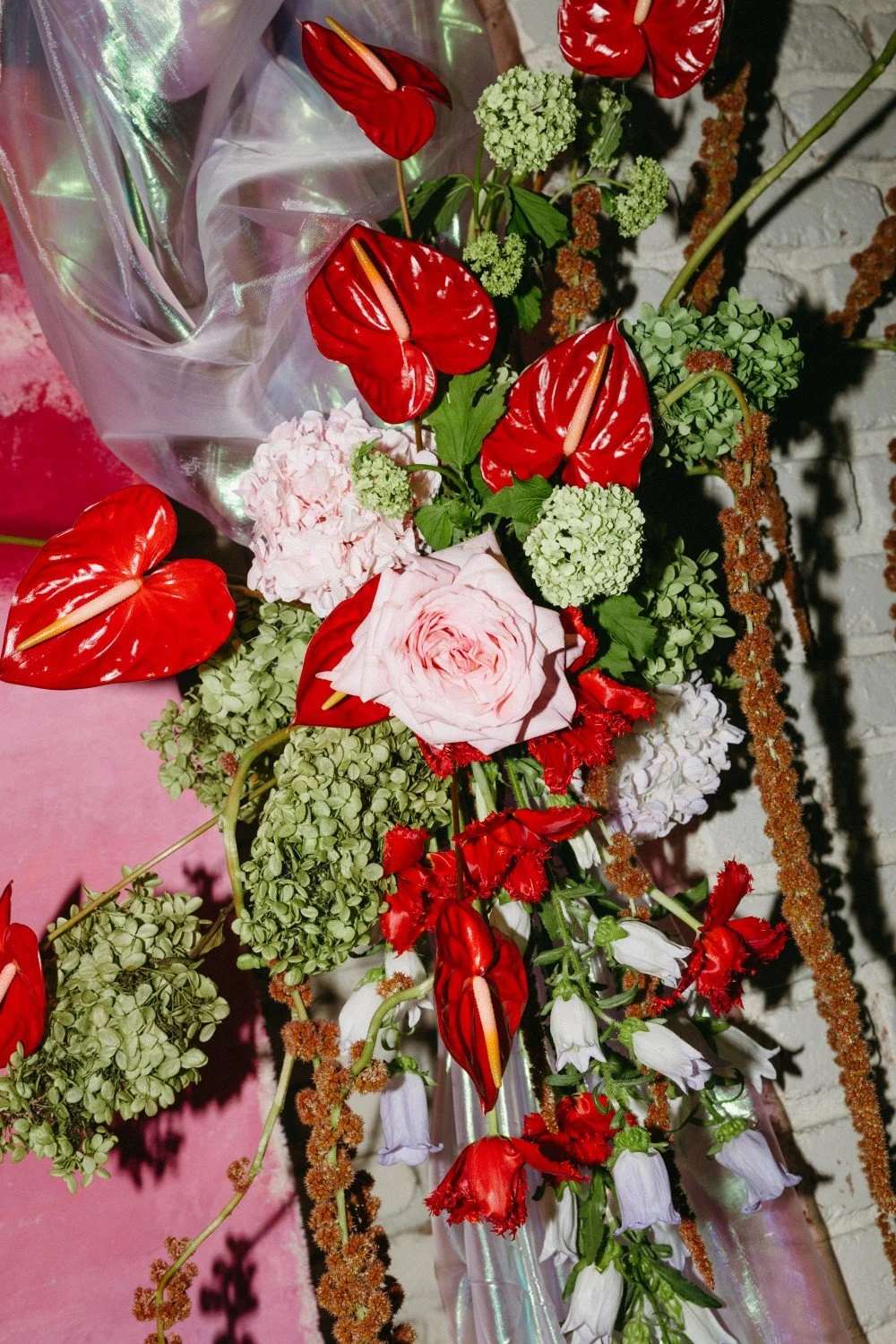 Colorful flower arrangement with red anthuriums, pink roses, white hydrangeas, and greenery, wrapped in clear cellophane.
