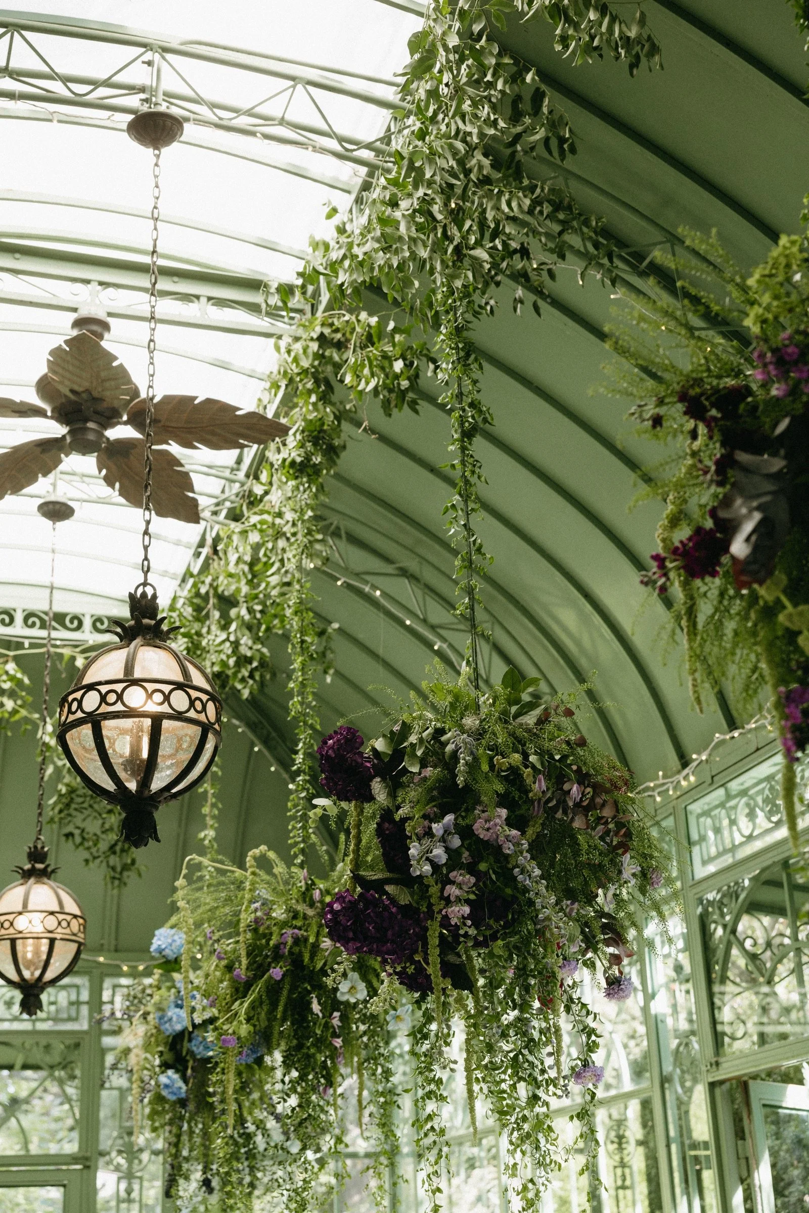 Hanging floral arrangements with greenery and purple, white, and blue flowers in a greenhouse with green metal framework and hanging lamps.
