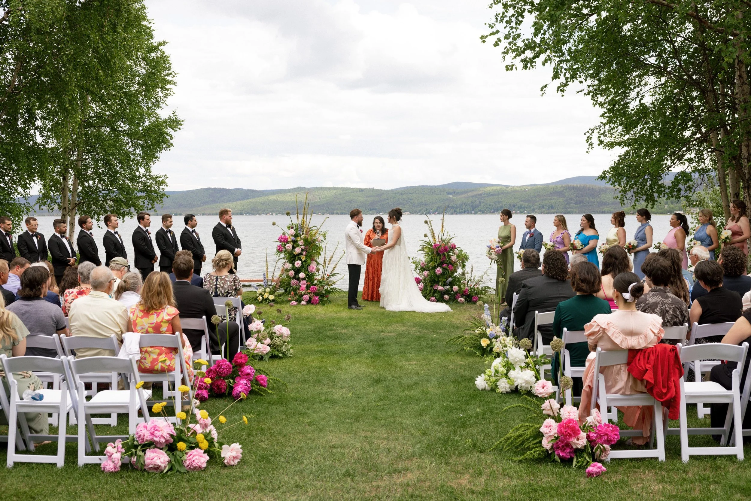 A wedding ceremony outdoors by a lake, with the bride and groom exchanging vows in front of friends and family, surrounded by floral decorations and lush green trees.