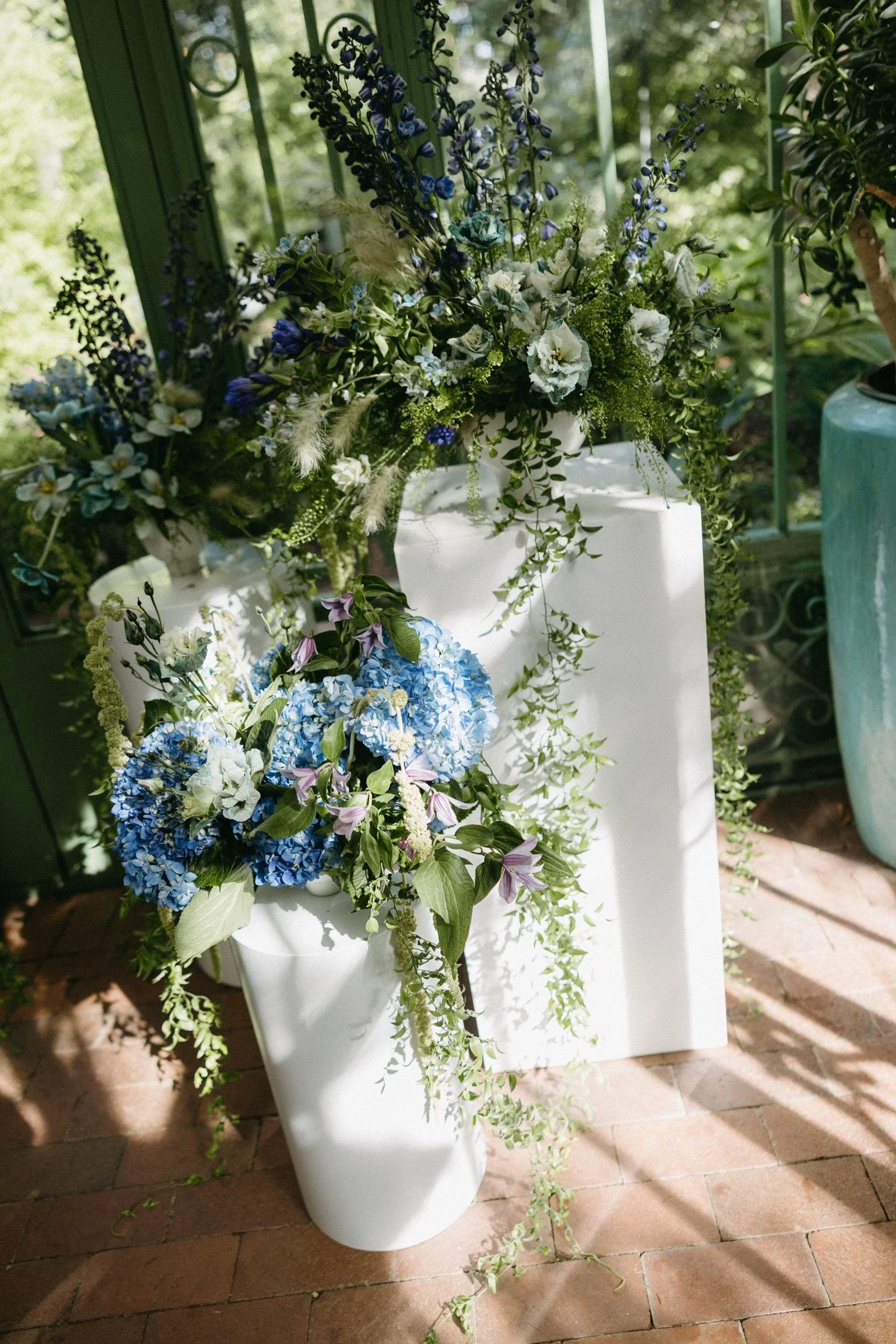 Elegant floral display with blue hydrangeas, white and purple flowers, and green foliage arranged in white vases, set against a green garden backdrop.