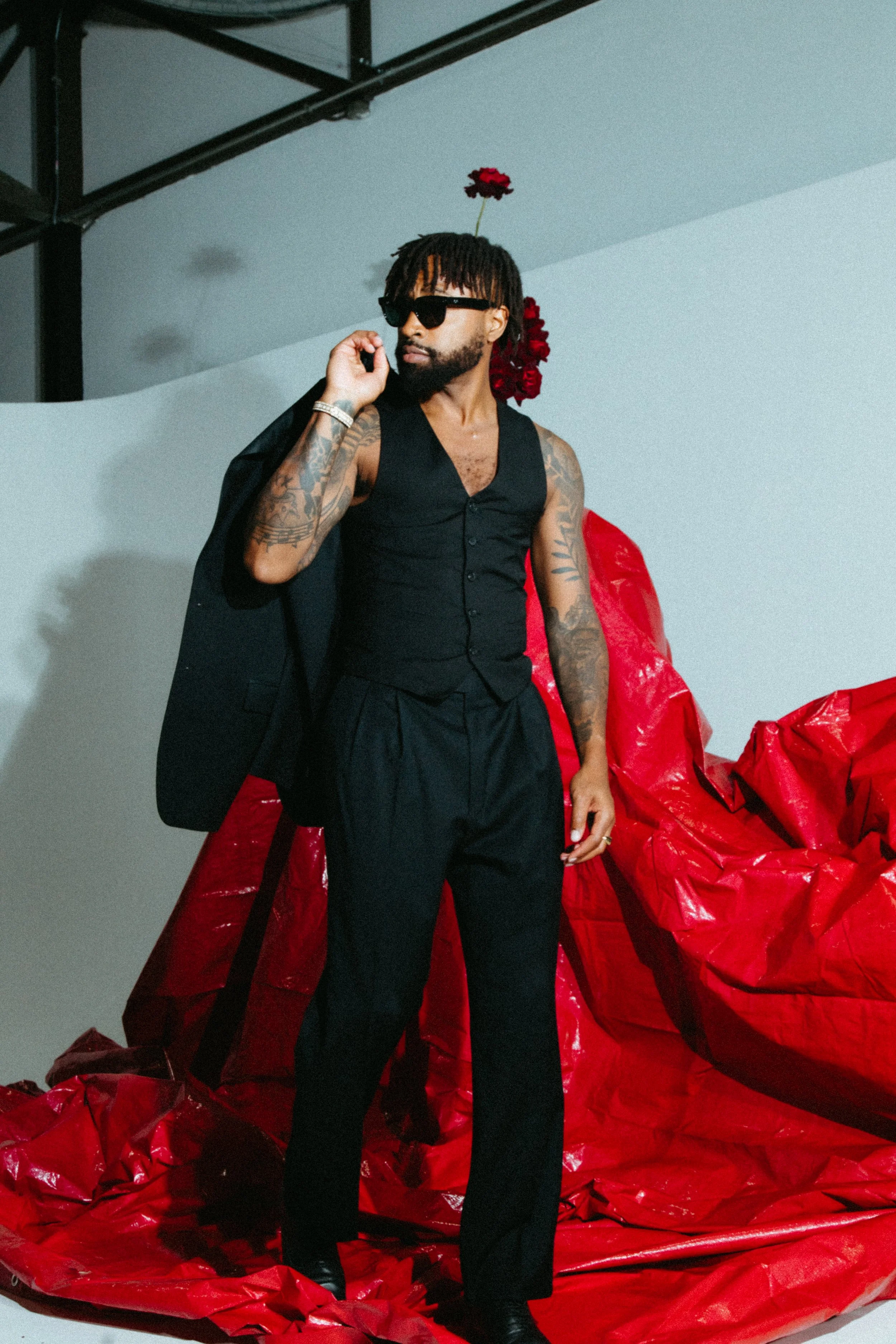 a Black male model in a dark suit against a background of white with a red backdrop and red carnations and roses