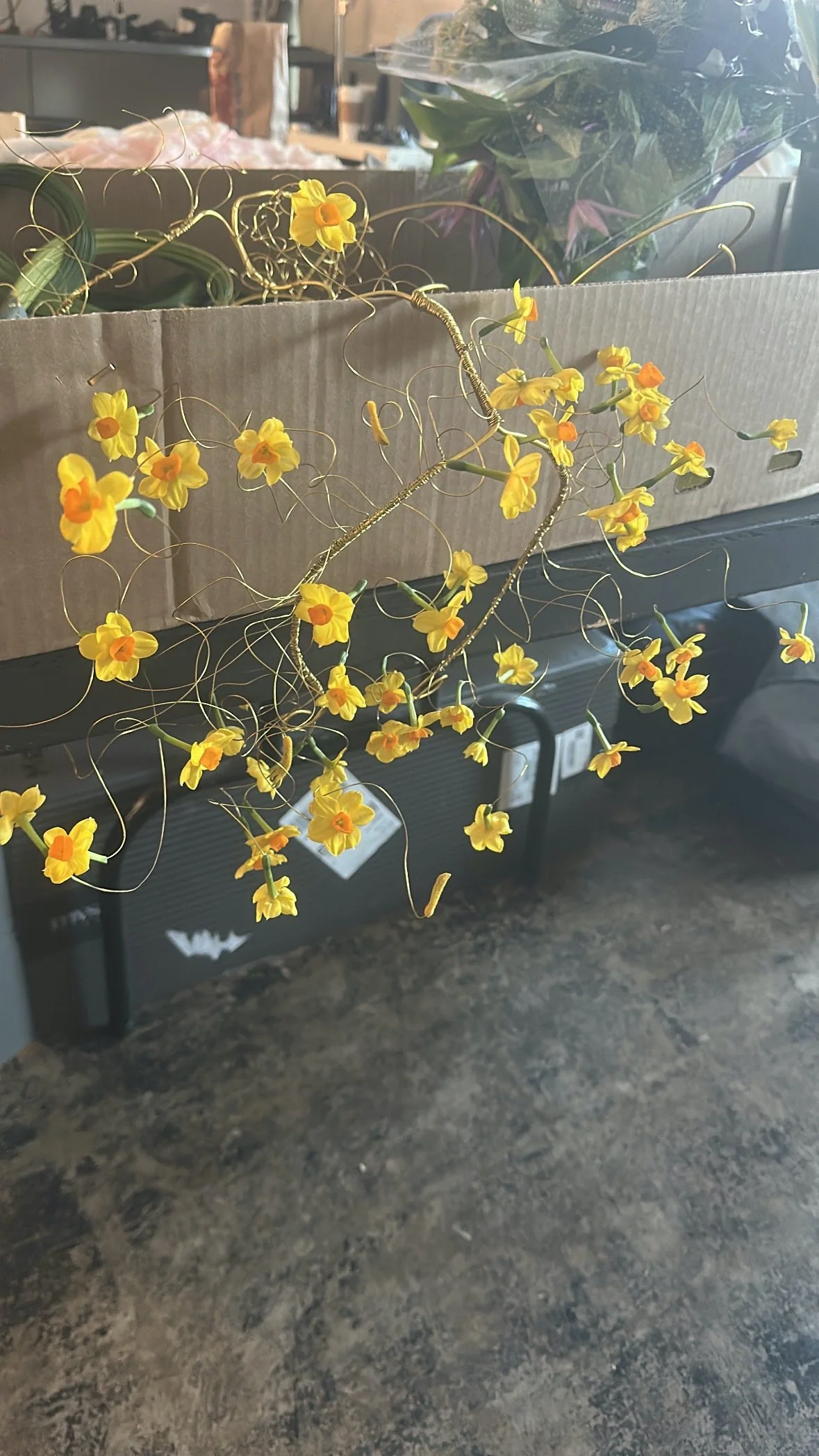A yellow artificial flower arrangement with curly wires, placed in front of a cardboard box, on a dark surface, with boxes and plants blurred in the background.