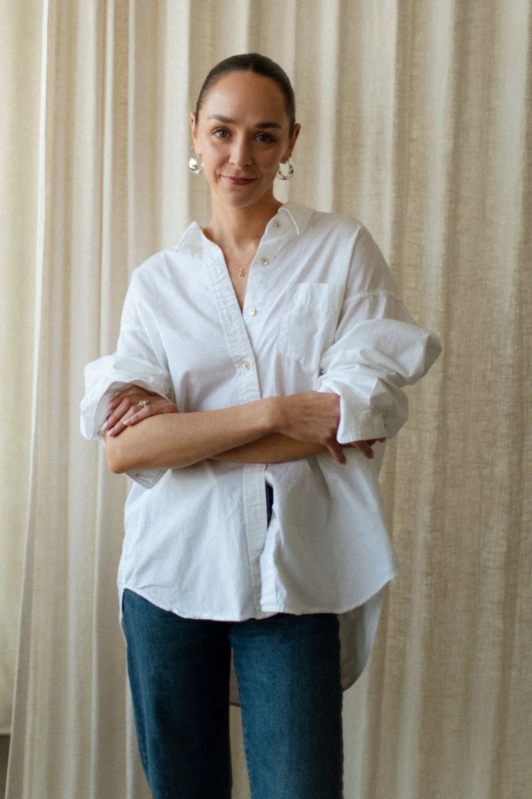 Woman with dark hair tied in a bun wearing a white button-up shirt with rolled-up sleeves and blue jeans, standing with arms crossed in front of beige curtains.