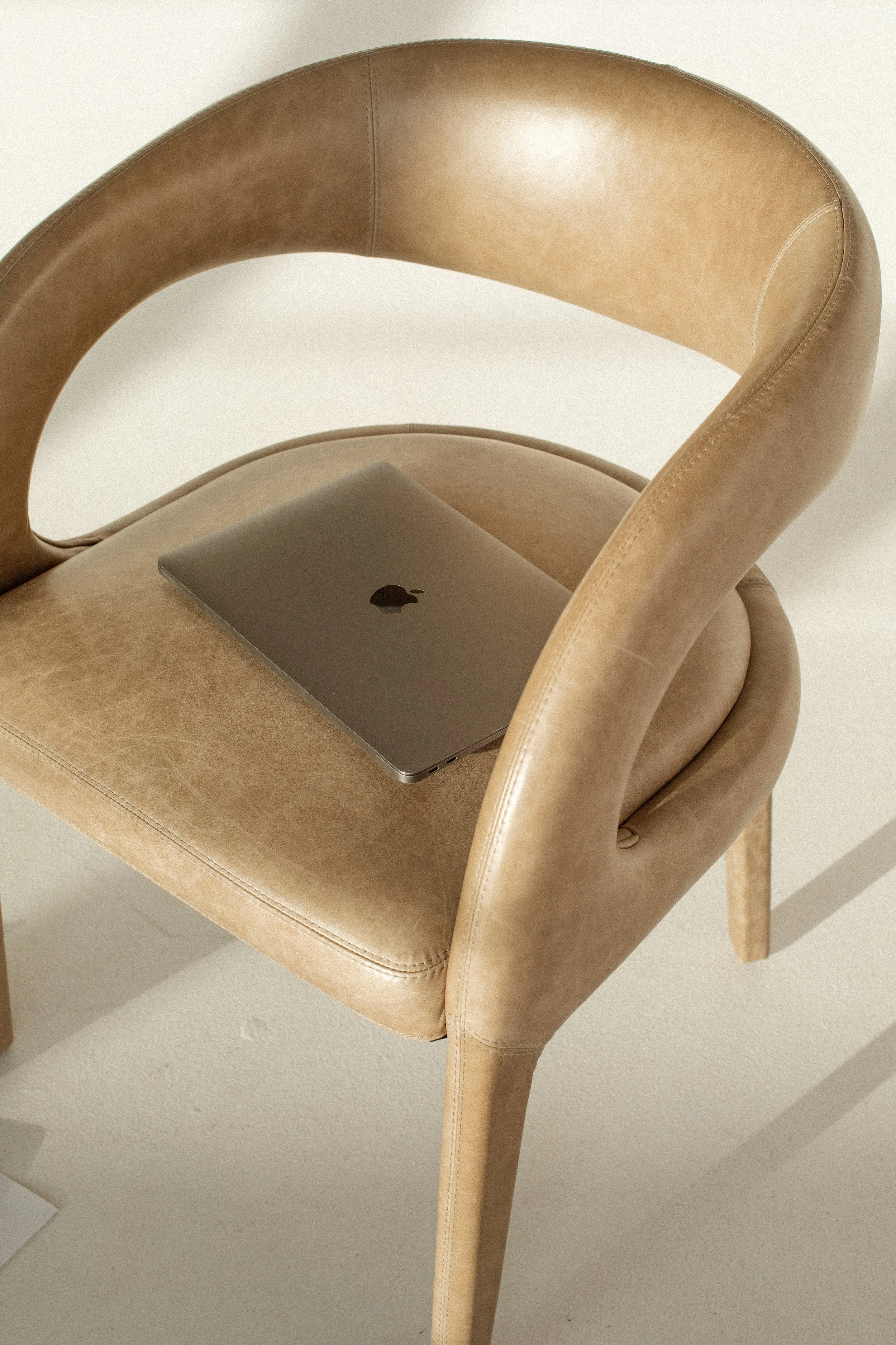 A beige leather chair with a closed MacBook laptop resting on its seat. The background is plain and off-white.