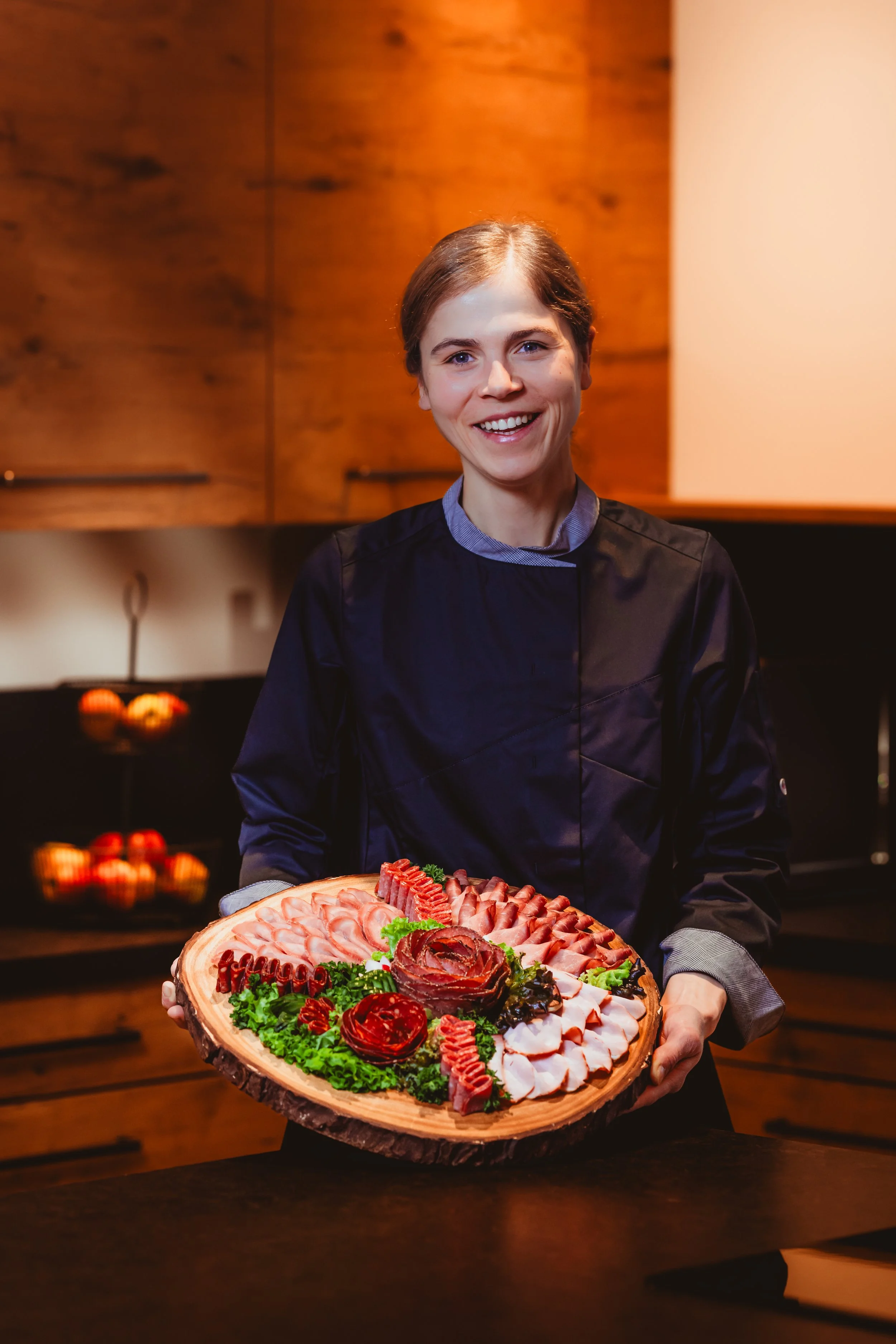 Eine Frau in Kochkleidung hält eine große Holzplatte mit verschiedenen Fleisch- und Wurstsorten, dekorativ auf grünen Blättern präsentiert.