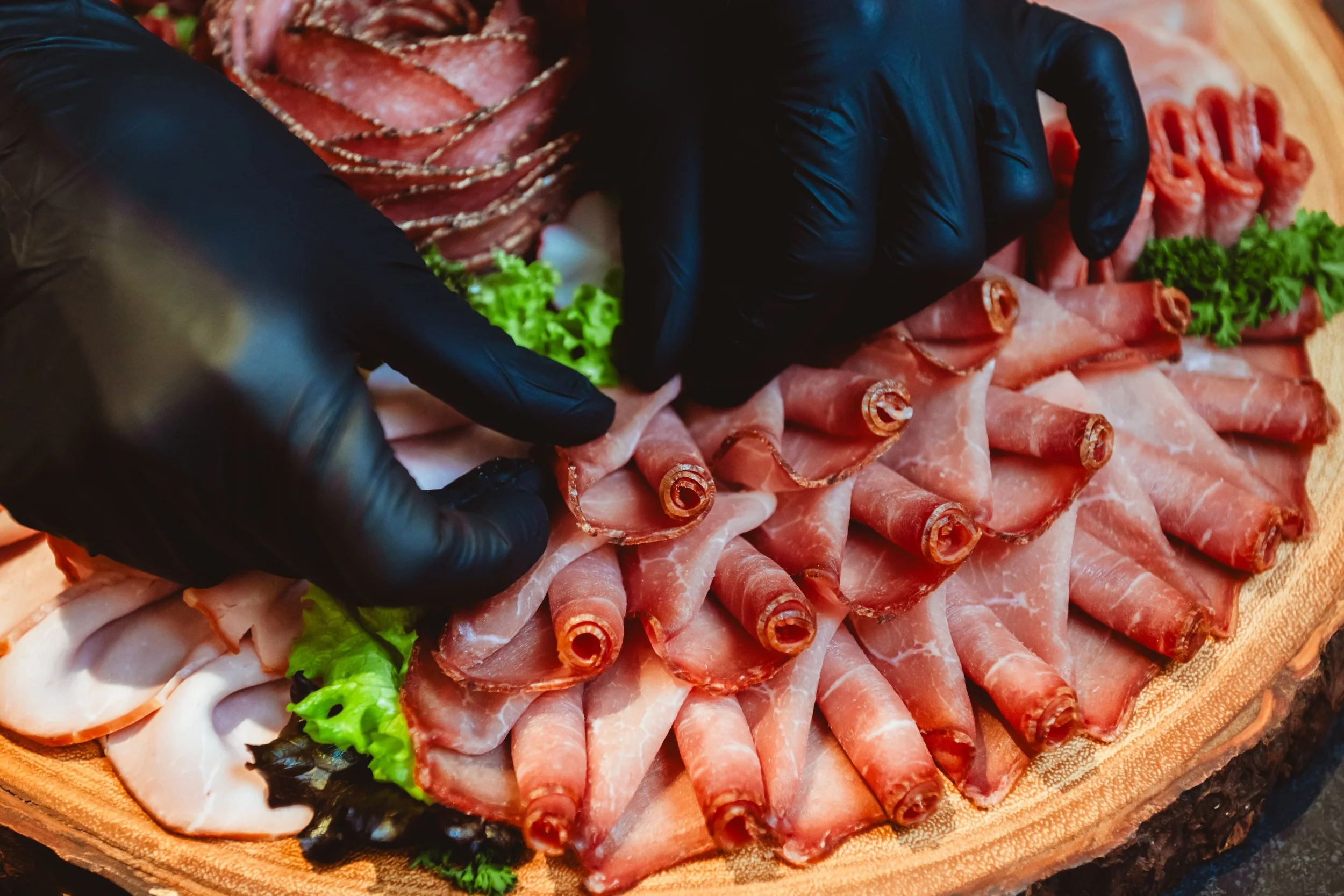Nahaufnahme von Händen in schwarzen Handschuhen, die Schinken- und Fleischröllchen auf einer Holzplatte arrangieren, umgeben von Salatblättern und weiteren Fleischsorten.