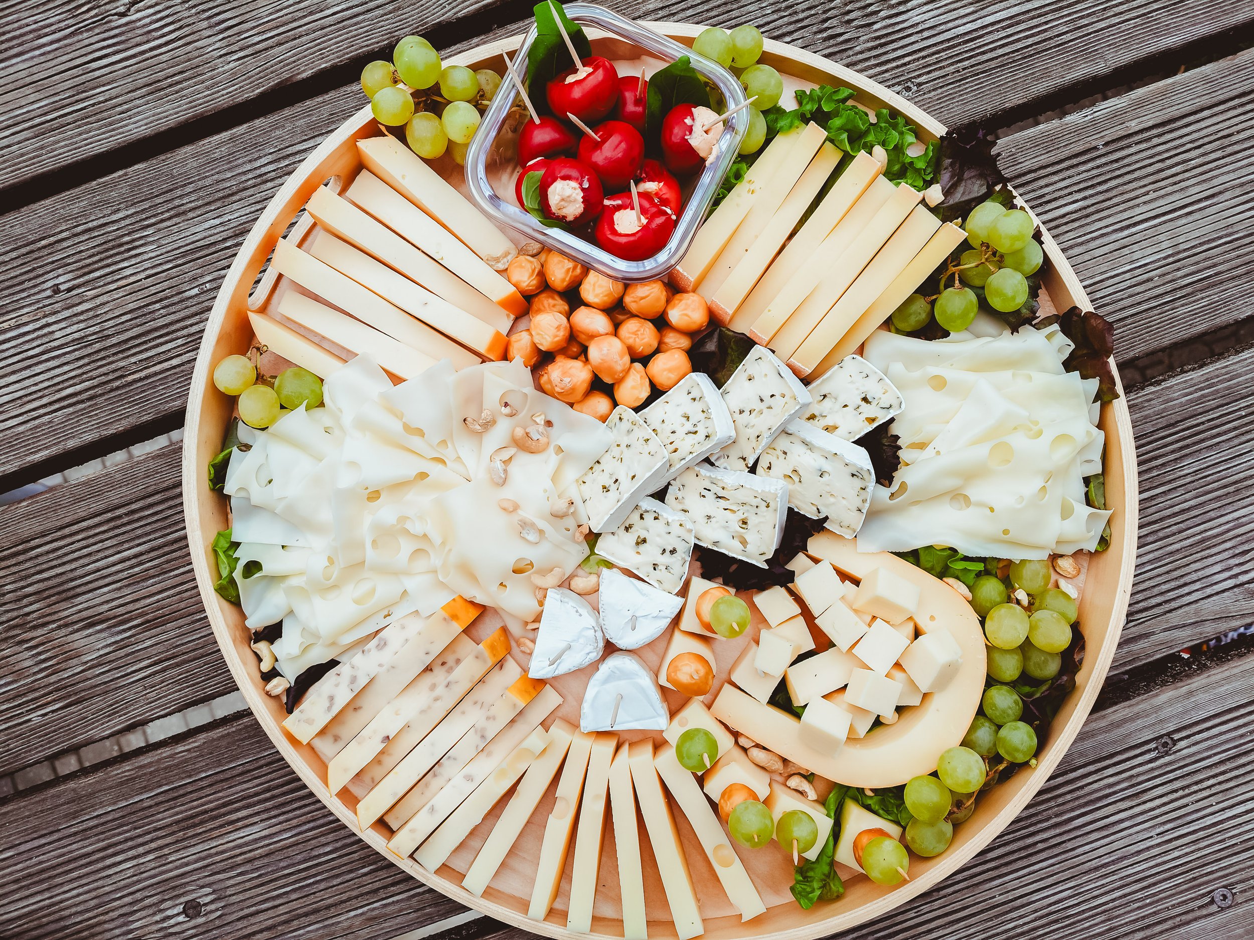 Käseplatte mit verschiedenen Käsesorten, Trauben, Cherrytomaten, Nüssen und einem kleinen Schälchen mit eingelegten roten Paprikaschoten.