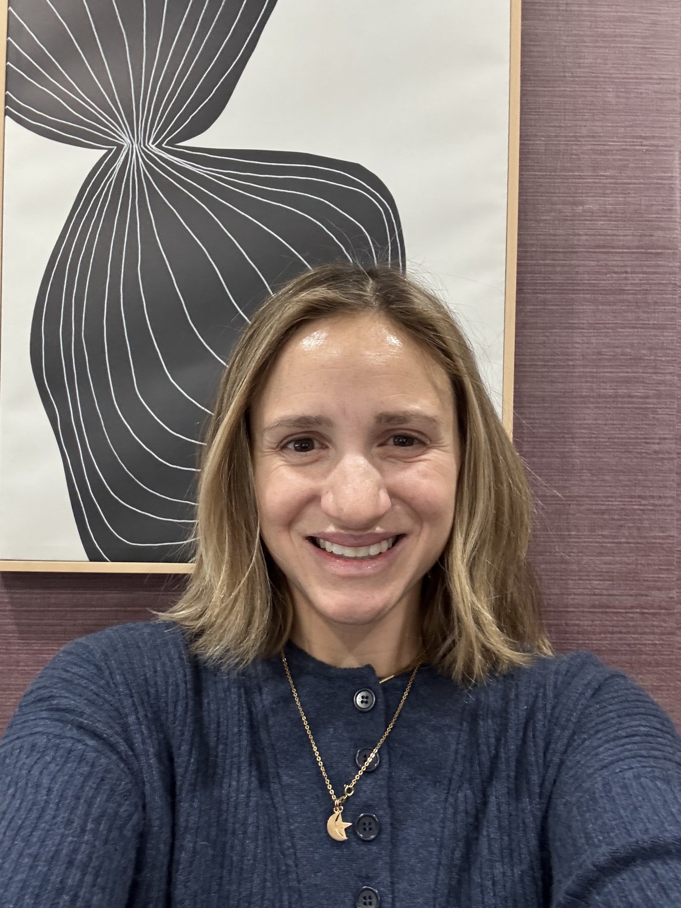 A woman with shoulder-length light brown hair taking a selfie indoors, smiling, wearing a navy blue button-up top and a gold necklace with a moon charm, with an abstract black and white line art poster and textured purple wall in the background.