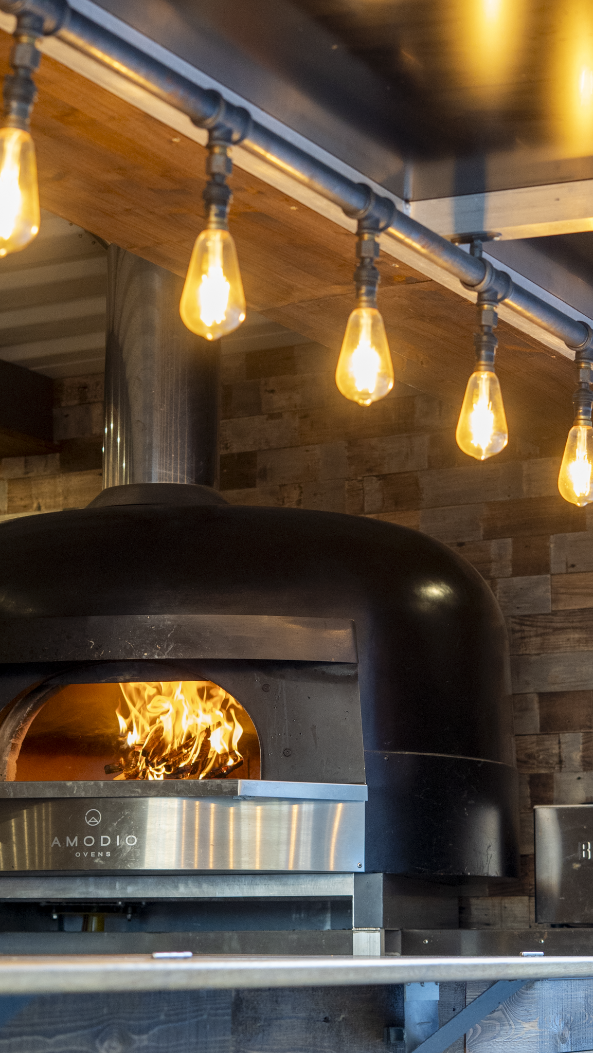 A modern wood-fired oven with a fire inside, situated below a wooden counter with hanging light bulbs.