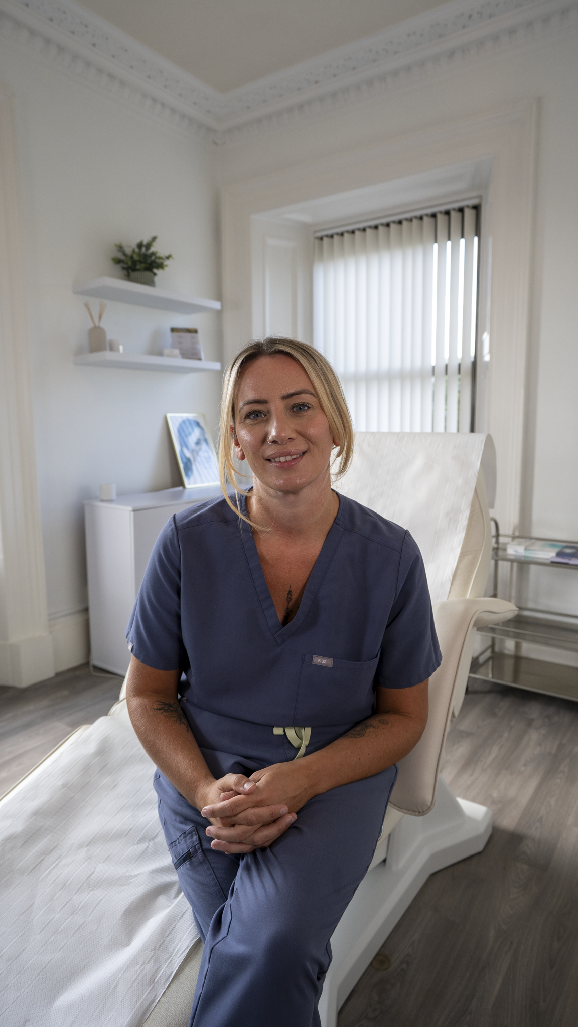 A female healthcare professional with blonde hair, wearing blue scrubs, sitting on the edge of an exam bed, smiling in a medical office or clinic room with white walls, a window with vertical blinds, and shelves with decor.
