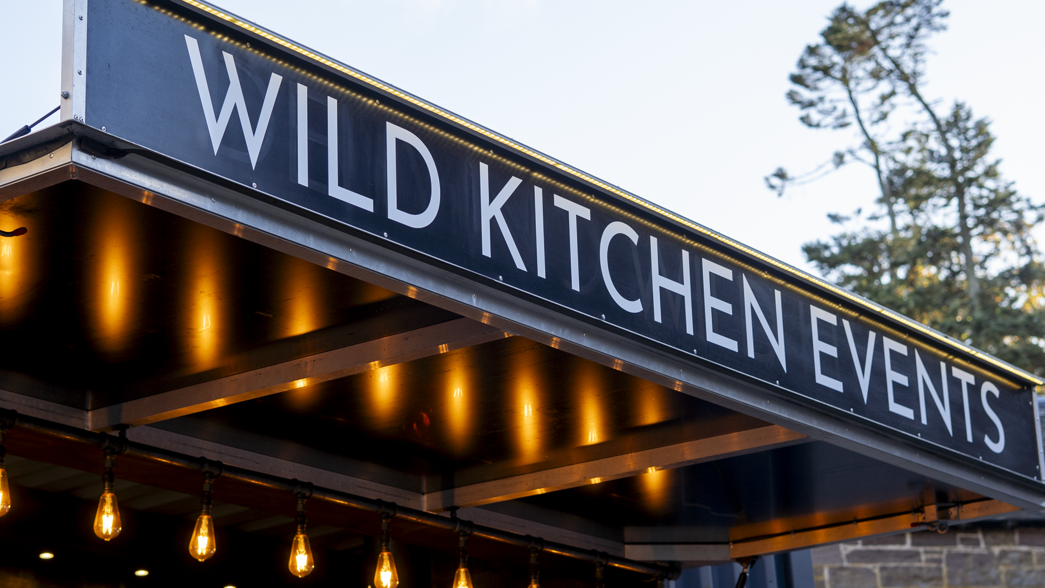 Sign that reads 'WILD KITCHEN EVENTS' above an outdoor event space with hanging warm-colored lights and trees in the background.