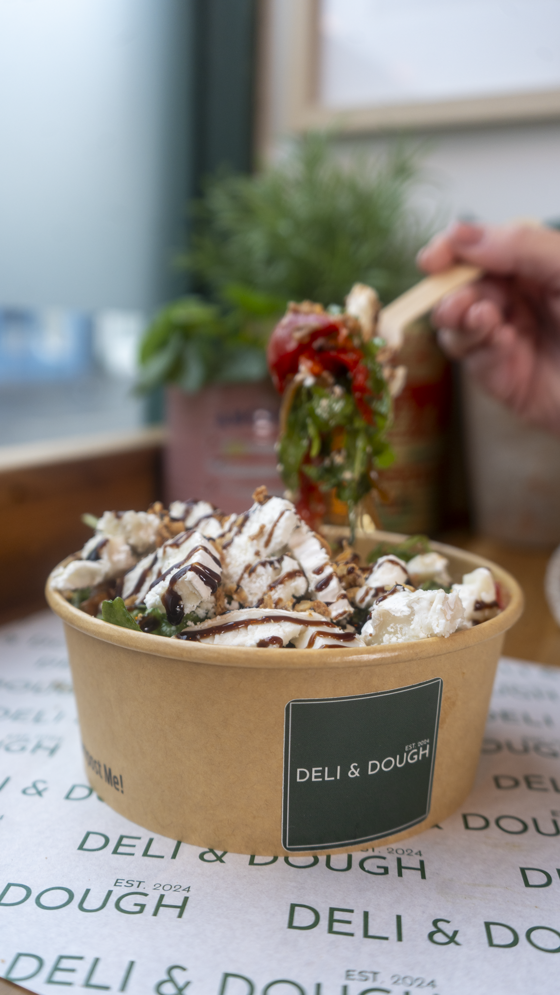 Close-up of a dessert bowl with ice cream topped with chocolate drizzle, whipped cream, chopped nuts, and a hand holding a spoon with a piece of cake and strawberries over it.