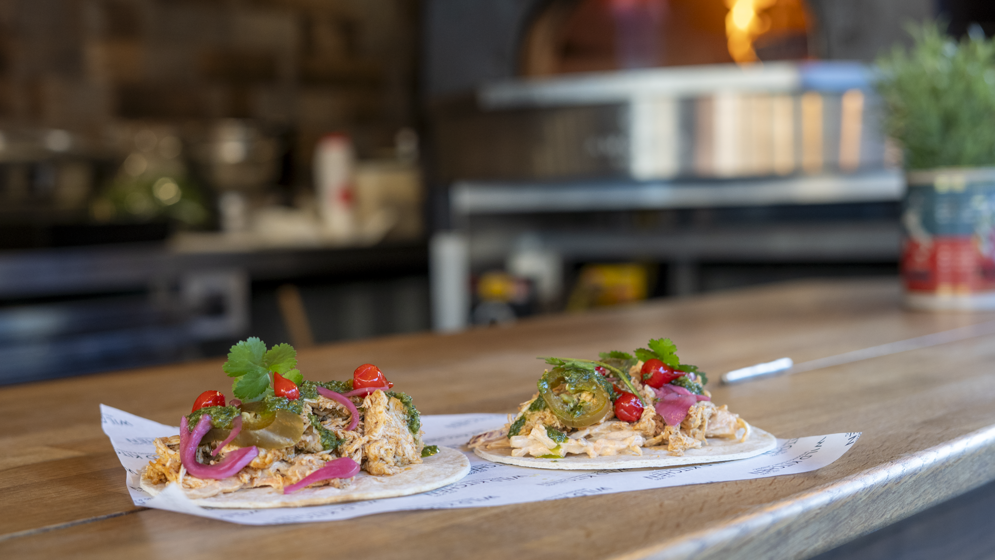 Two small tacos on a wooden counter, topped with cilantro, jalapeños, pickled red onions, and red peppers, with a blurry kitchen background.