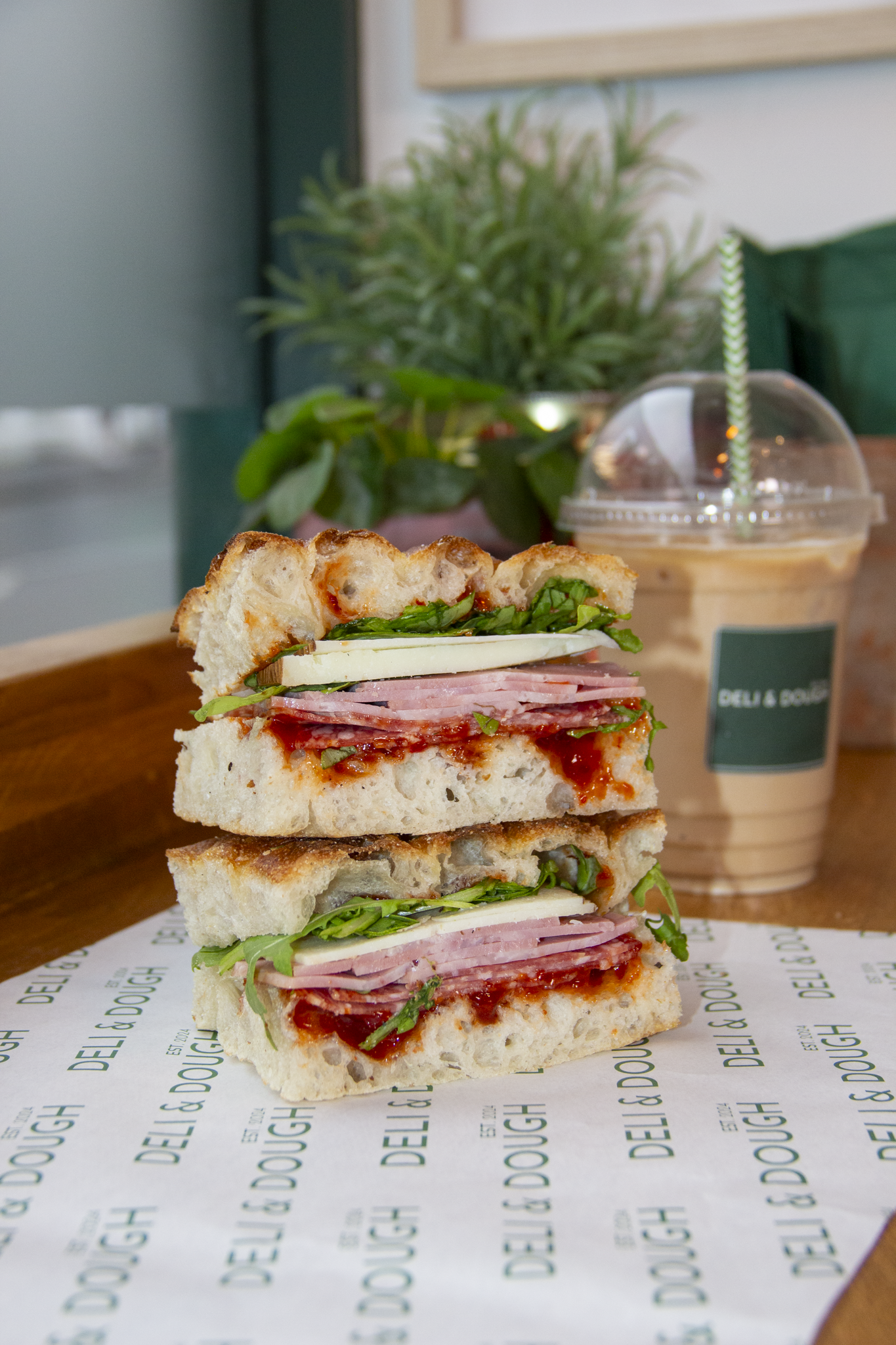 A sandwich with layers of ham, cheese, lettuce, tomato, and basil on focaccia bread. A cup of iced coffee with a straw is in the background, along with a green plant and a framed picture.