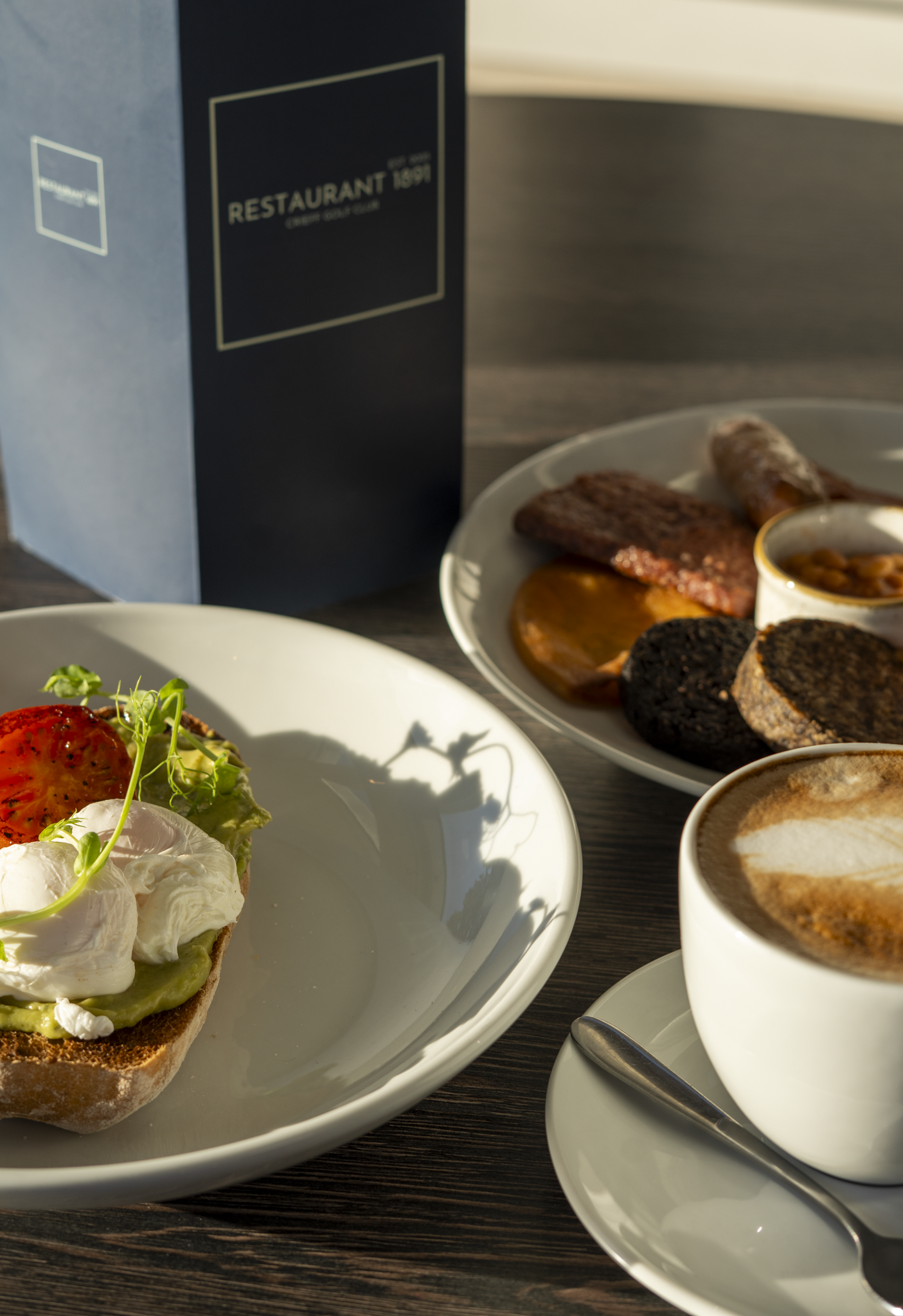 A breakfast setting with a plate of avocado toast topped with poached eggs and tomato slices, a cup of cappuccino with frothy foam and cinnamon, and a plate of assorted breakfast meats and dishes in a black and white box with the text 'RESTAURANT 1891'.