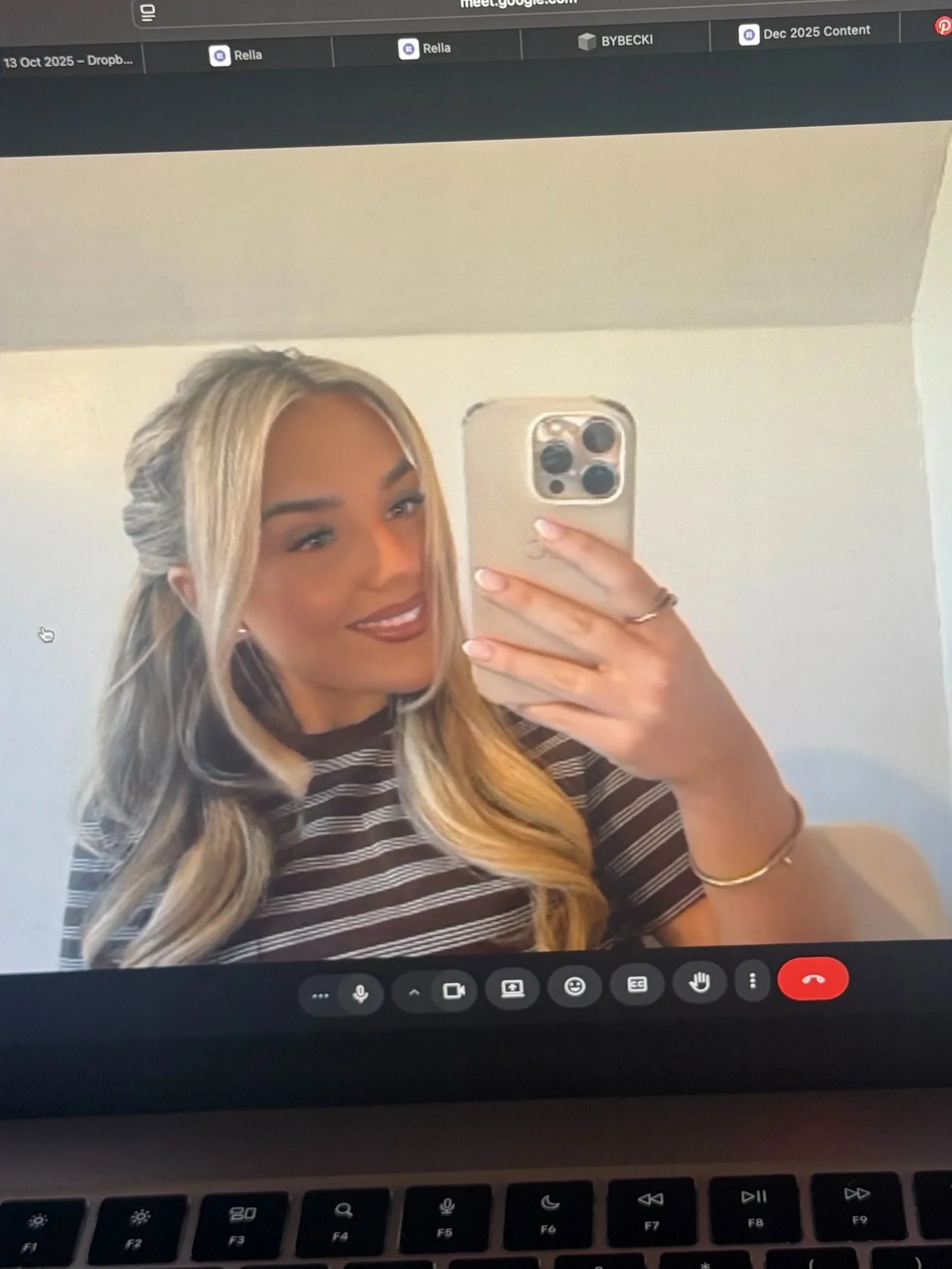 A woman taking a selfie during a video call on a computer, smiling, with blonde hair, wearing a striped shirt, and holding her phone.