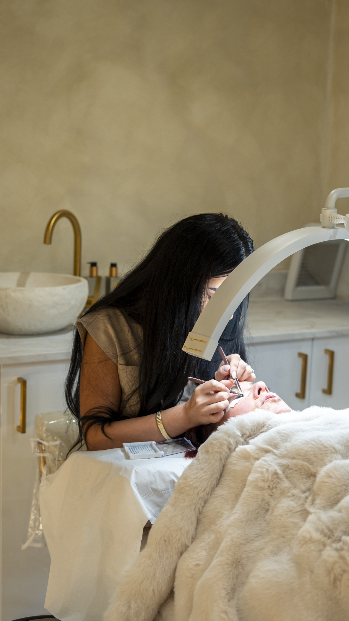 A woman receiving a cosmetic or dermatological treatment on her face in a clinical setting, with another woman performing the procedure under a magnifying lamp.