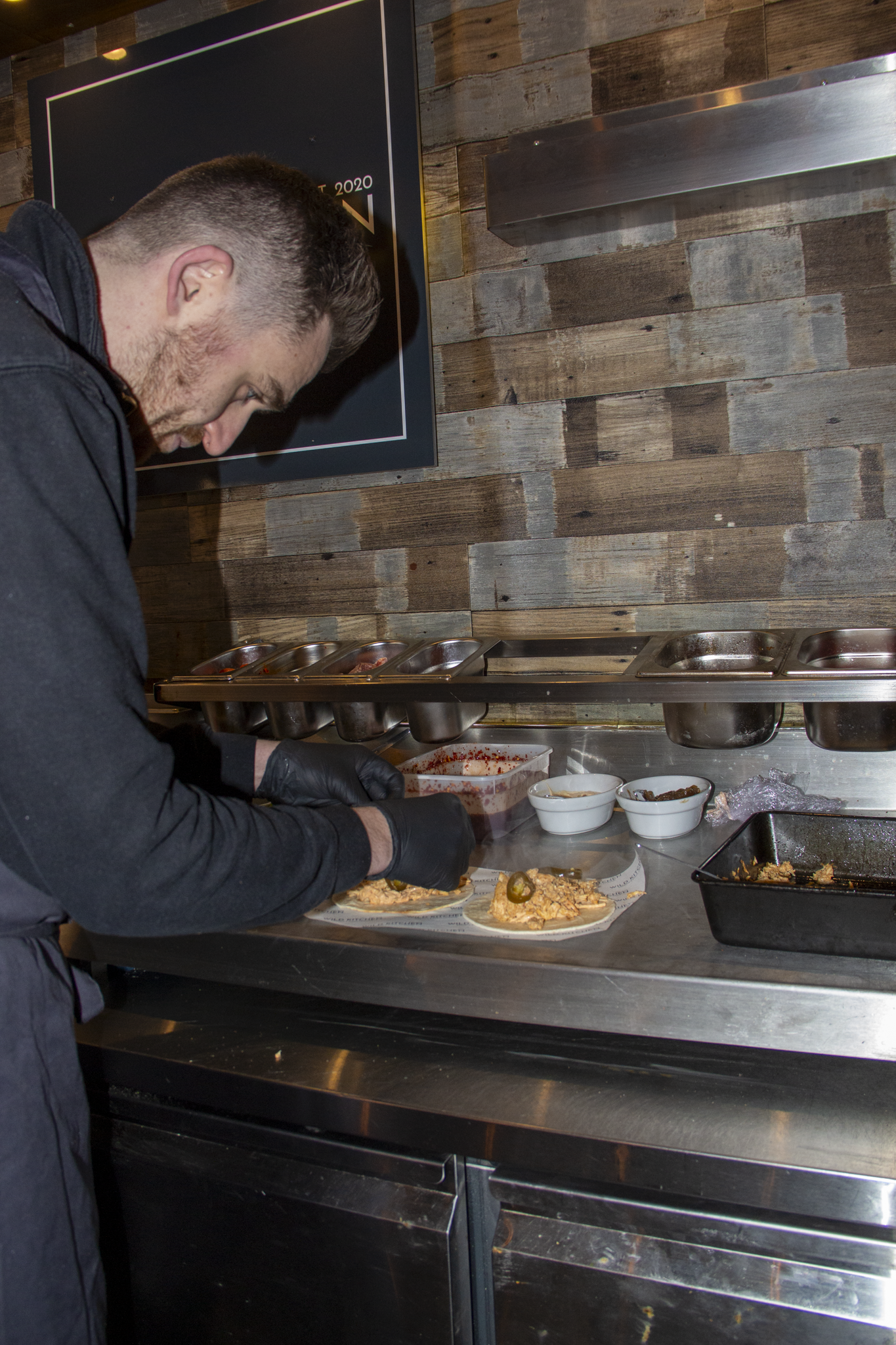 A man wearing black gloves preparing food on a stainless steel counter inside a restaurant kitchen, with metal trays and bowls of ingredients. The background features a wooden wall with a framed poster.