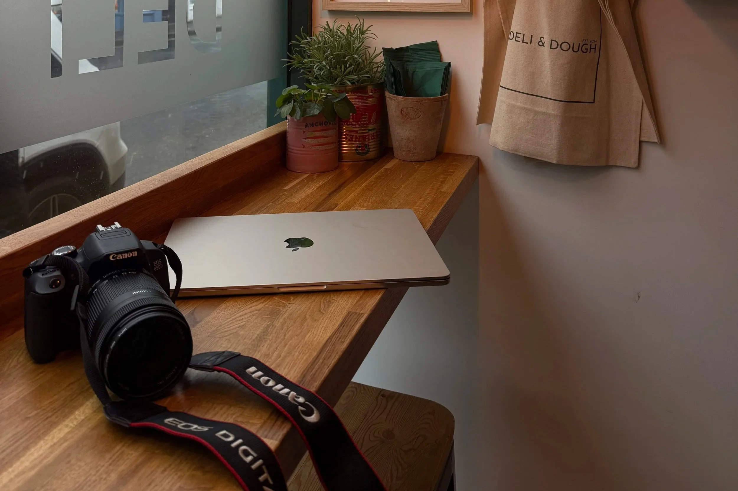 A wooden counter with a Canon camera and a closed Apple MacBook laptop on it, along with a small potted plant, a can with a plant, and another container with green napkins, near a window with partly frosted glass, and a beige wall with a hanging rolled-up canvas print.