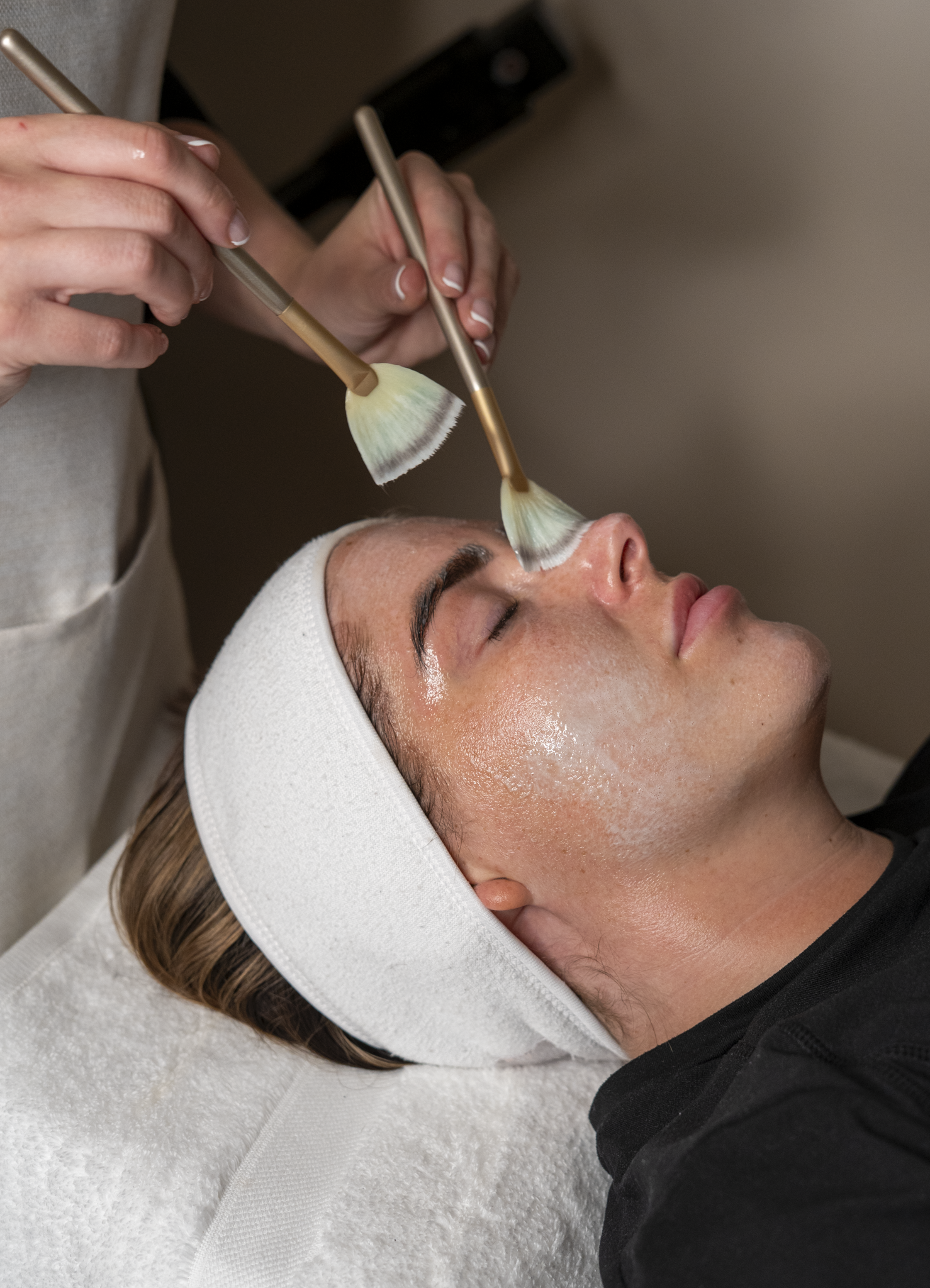 A woman receiving a facial treatment at a spa, with a cosmetologist applying skincare products using two brushes on her face.