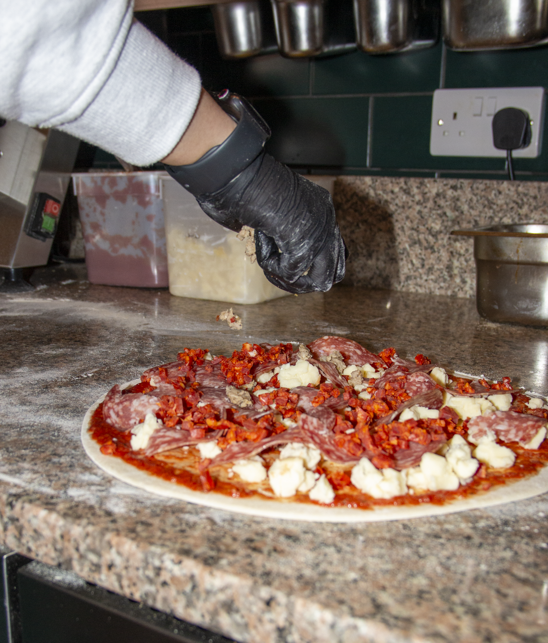 Person wearing black gloves preparing a pizza with various toppings on a granite countertop.