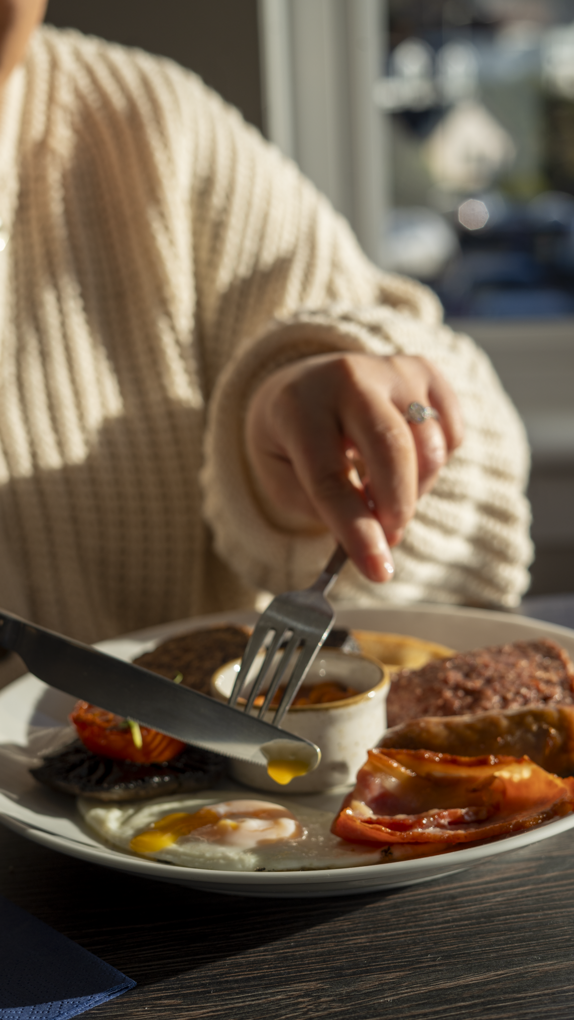 A person in a cream-colored, chunky knit sweater is cutting into a breakfast plate that includes eggs, bacon, sausages, and grilled vegetables.