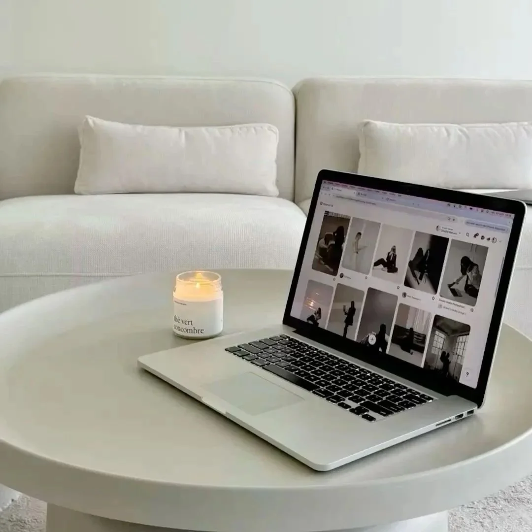 A white round coffee table with a lit candle and an open laptop displaying a grid of black and white images. In the background is a light-colored sofa with two matching pillows.