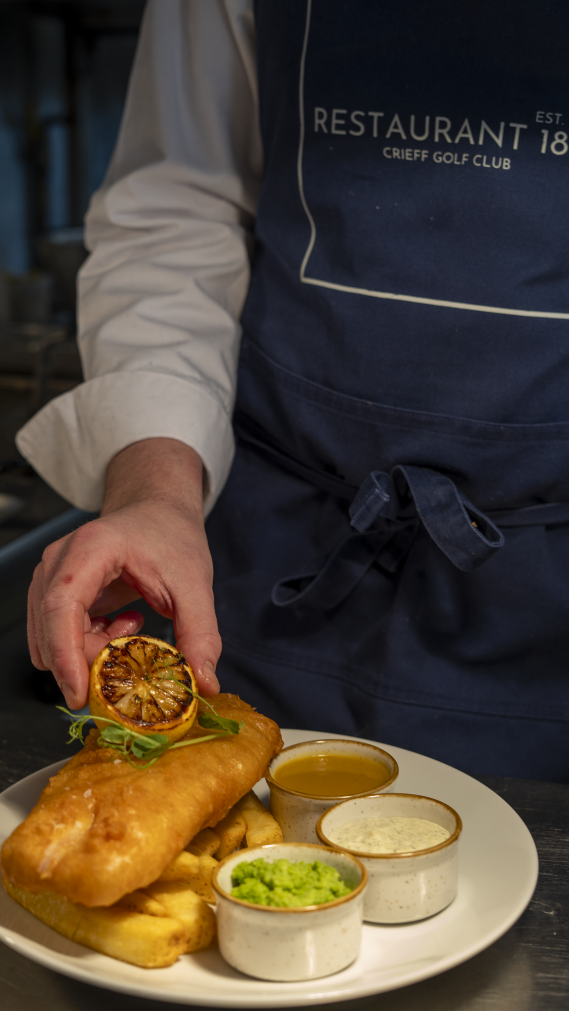 Chef garnishing battered fish and chips with a charred lemon half and microgreens, with three dipping sauces on the plate.
