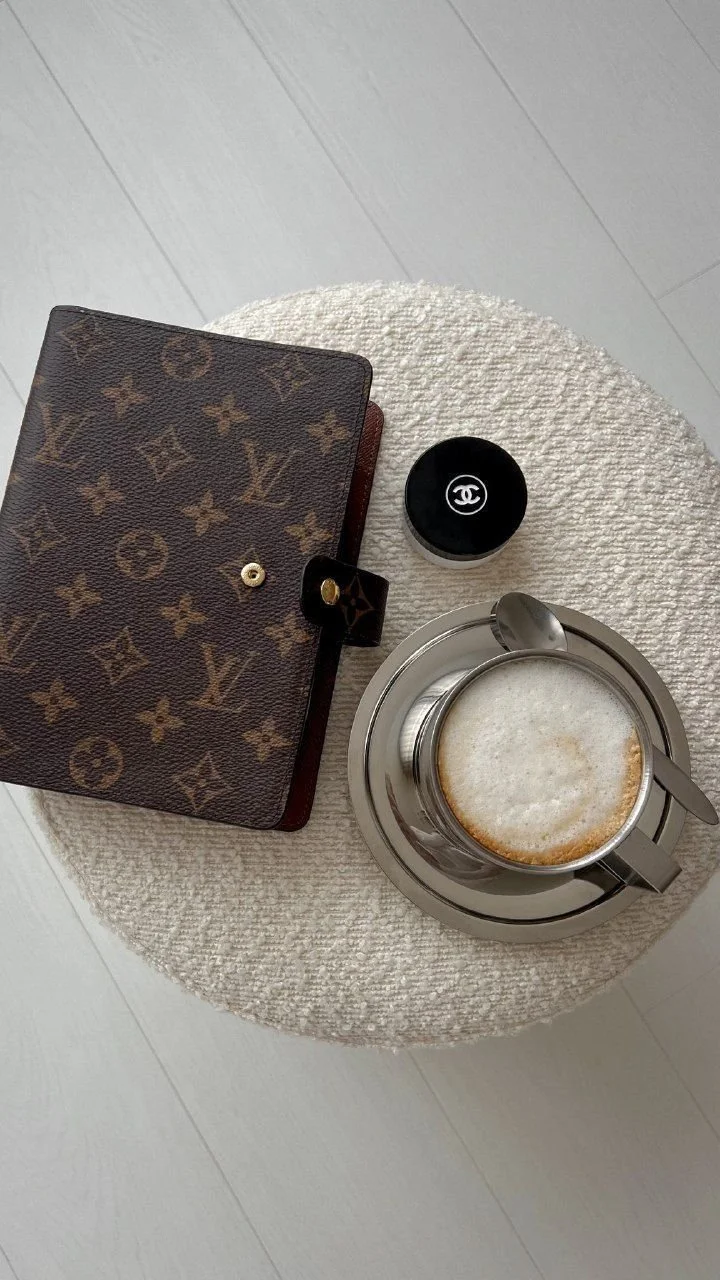 A round beige textured surface with a Louis Vuitton wallet, a small Chanel container, a glass of frothy cappuccino, and a spoon on a metal saucer.