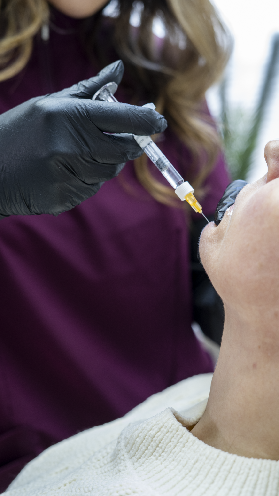 A person is receiving a dental shot from a healthcare professional wearing black gloves. The patient is lying back in a dental chair, with their mouth open, while the healthcare worker prepares to administer anesthesia or medication.