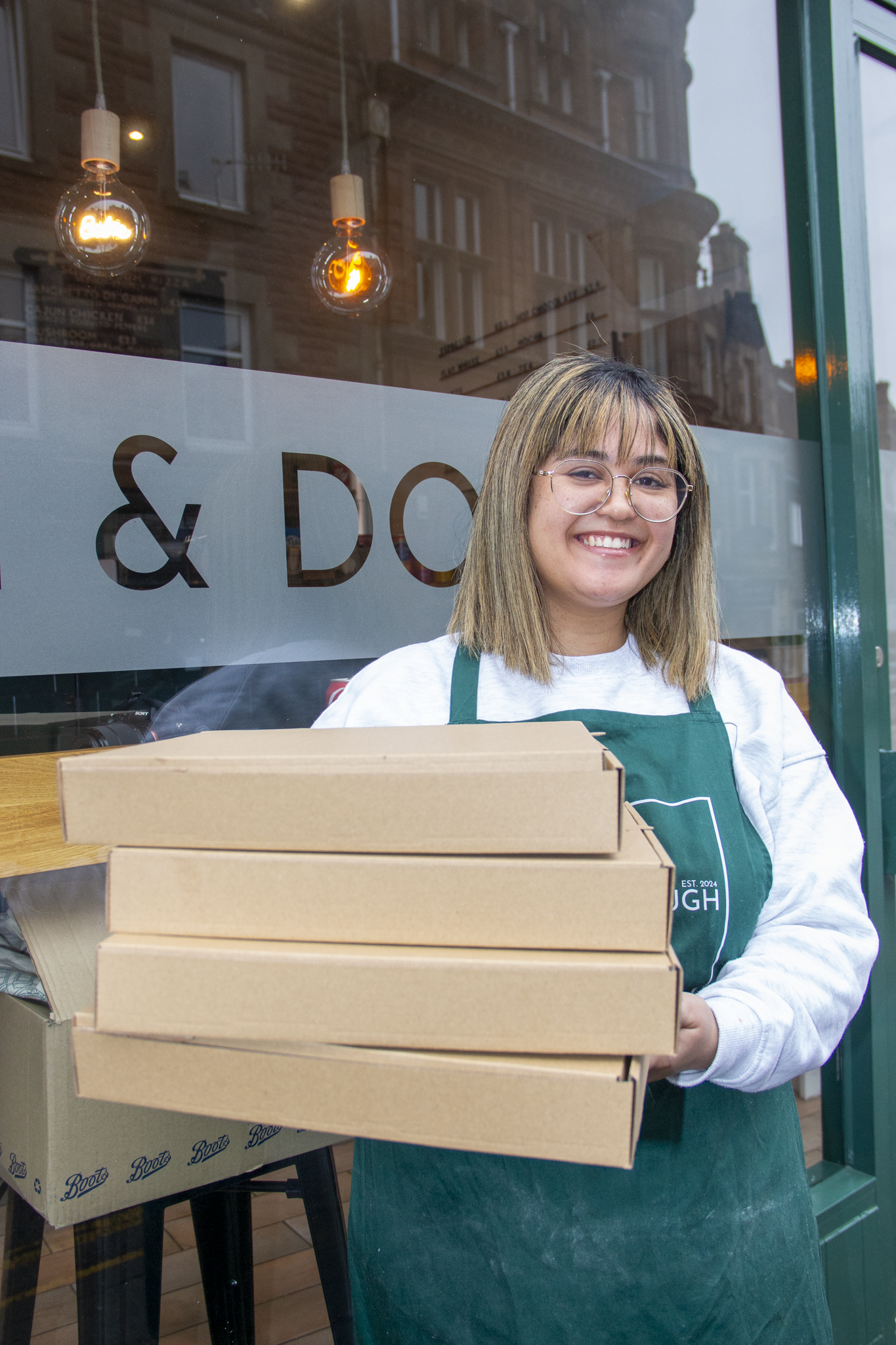 Smiling woman with glasses wearing a green apron holding four pizza boxes outside a storefront with a glass window.
