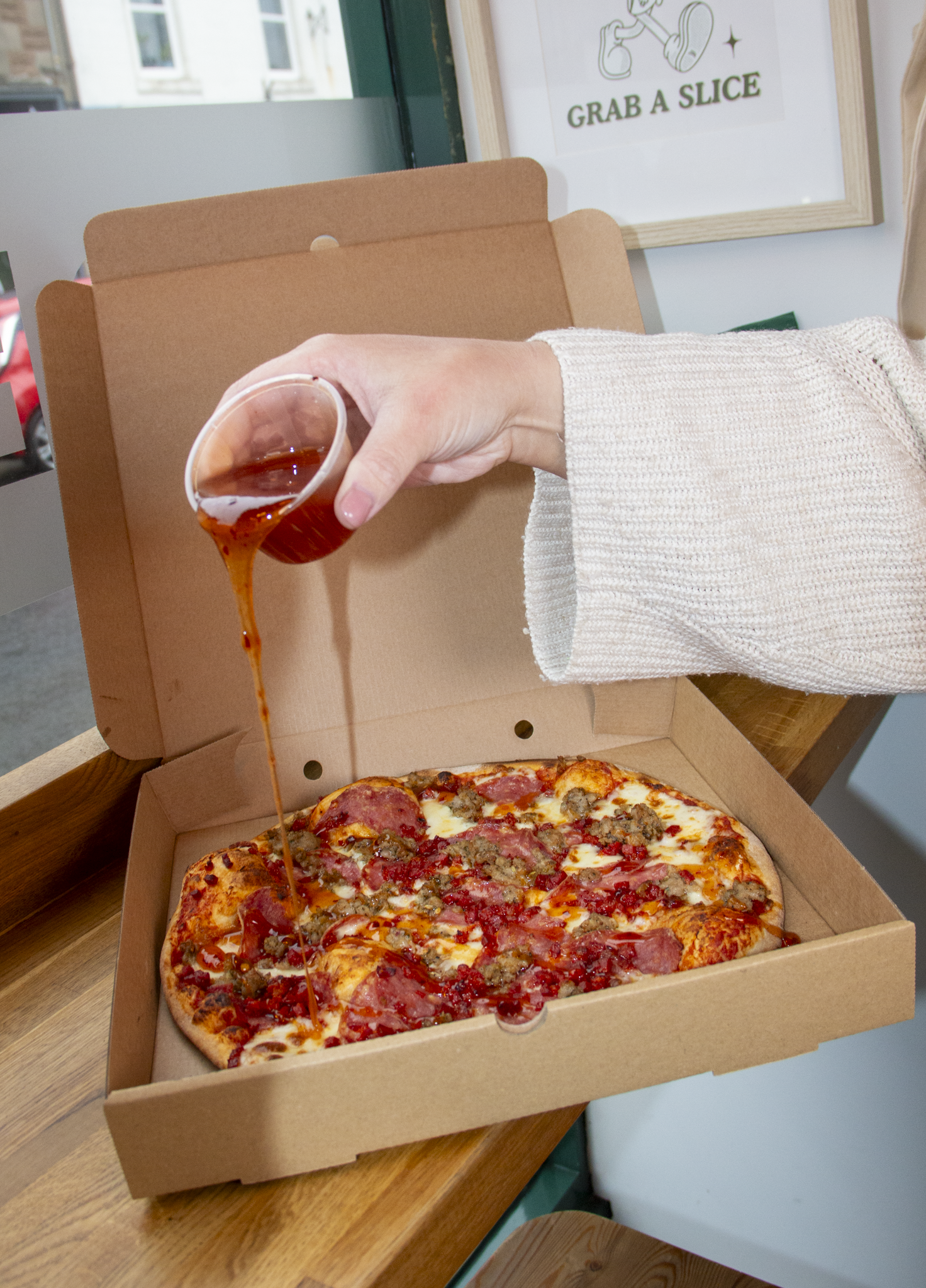 A person is pouring hot sauce or liquid into a pizza in an open cardboard box, with a sign in the background that says 'Grab a Slice'.
