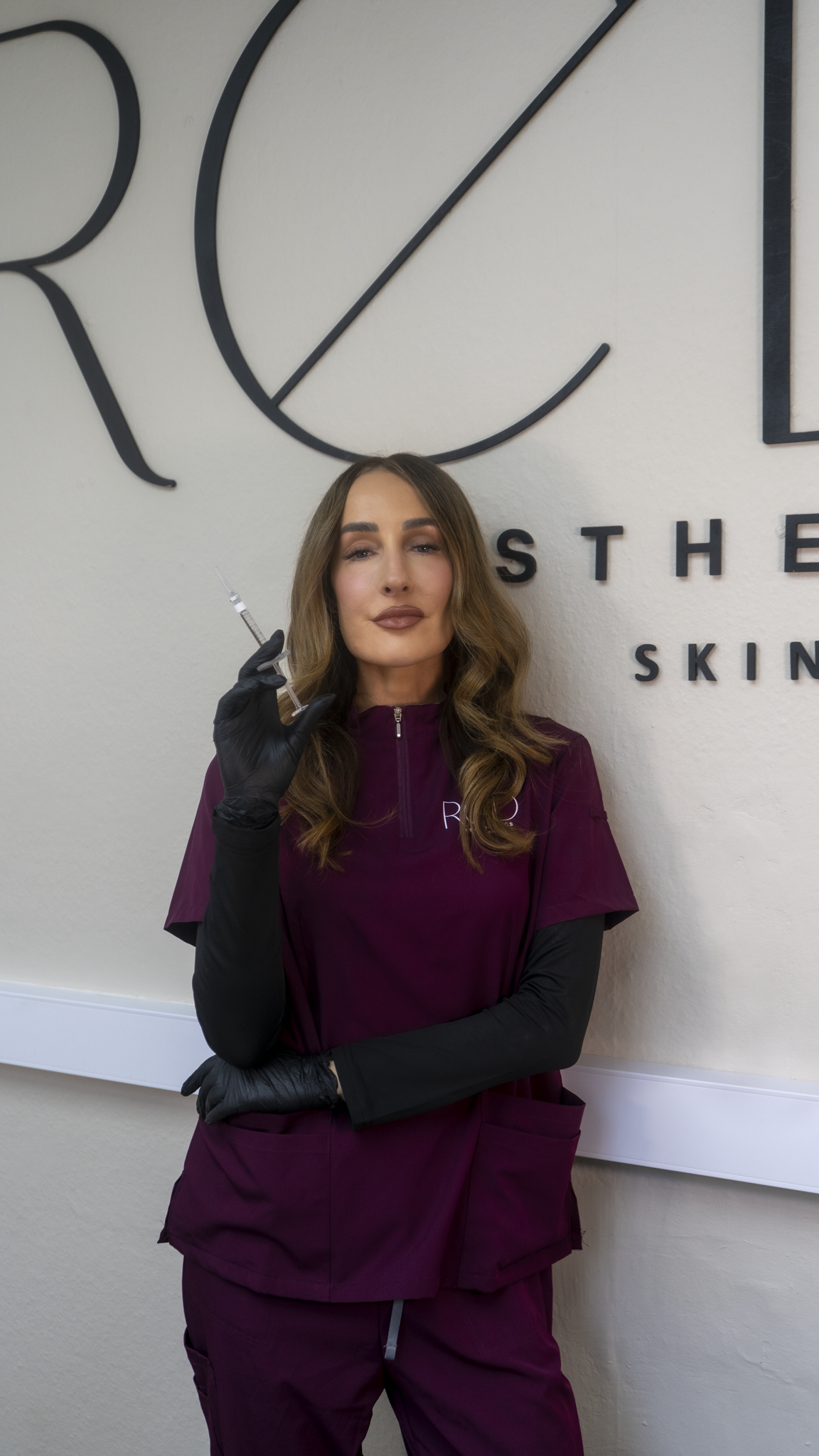 A woman in medical scrubs and black gloves holding a syringe in front of a wall with signage for a skincare clinic.