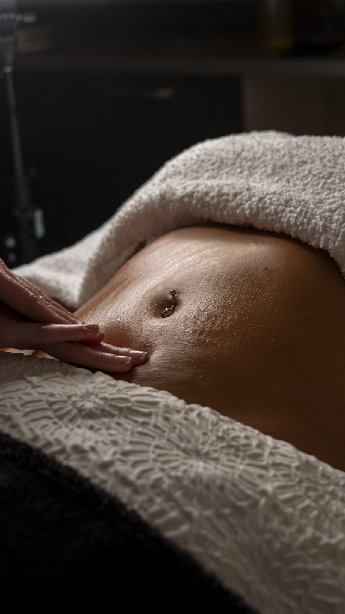Close-up of a person's bare abdomen with a belly button piercing, partially covered by a white textured towel or blanket, with a hand gently touching the lower abdomen.