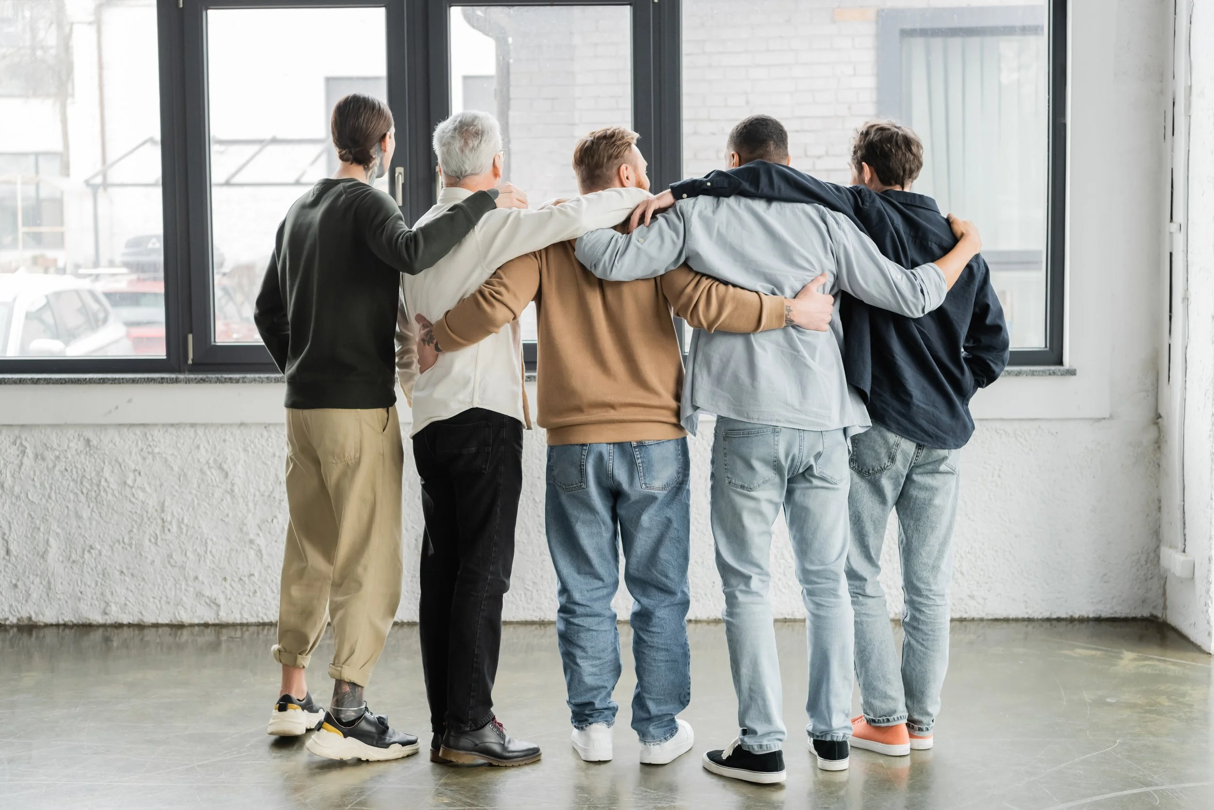 A diverse group of five people standing in a row with their arms around each other's shoulders, facing a large window in an indoor setting.