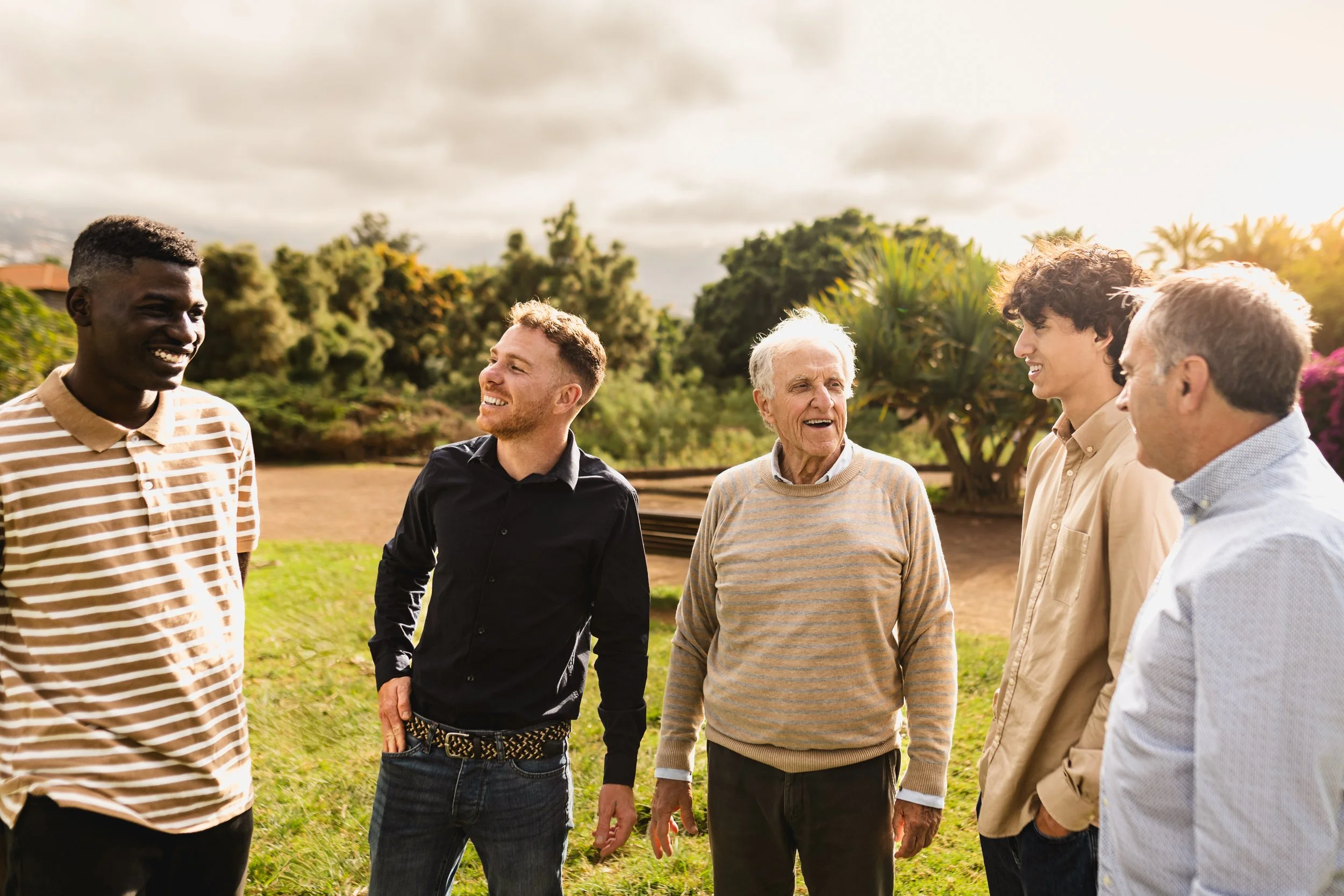 Group of five men standing outdoors in a park, engaged in conversation and smiling, with trees and cloudy sky in the background.