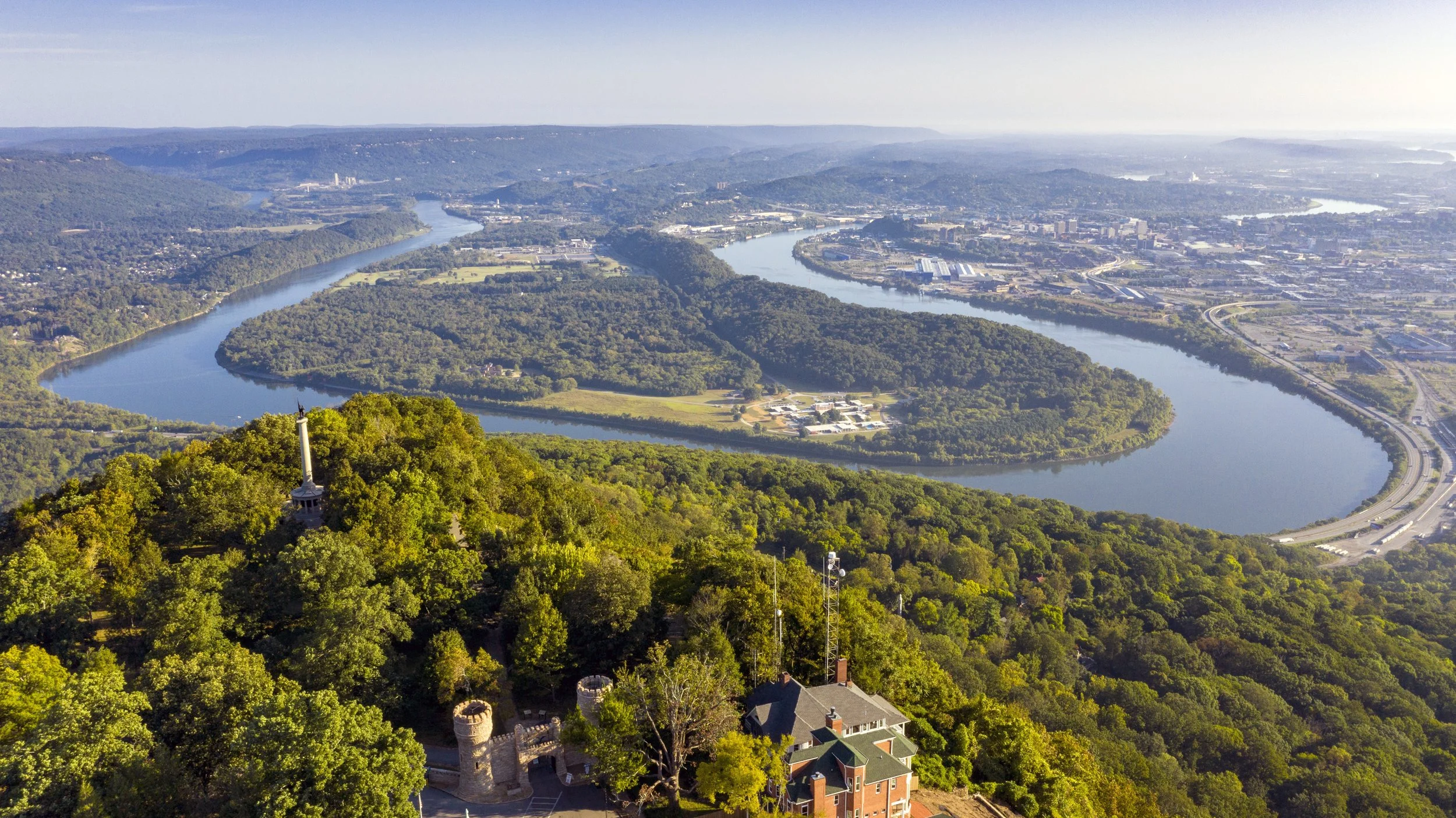 Aerial view of a winding river flowing through wooded hills and a city with industrial buildings in the distance, with a small castle and a church at the forefront.