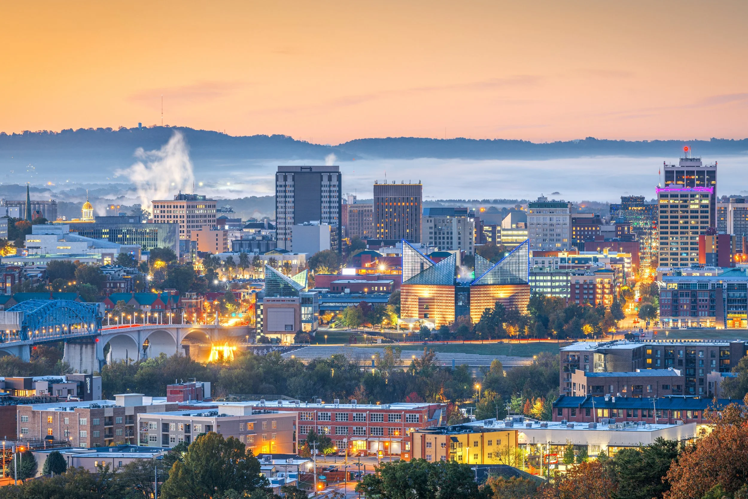 City skyline at dusk with buildings, a bridge, and hills in the background, some buildings are lit with colorful lights, and there is fog in the distance.
