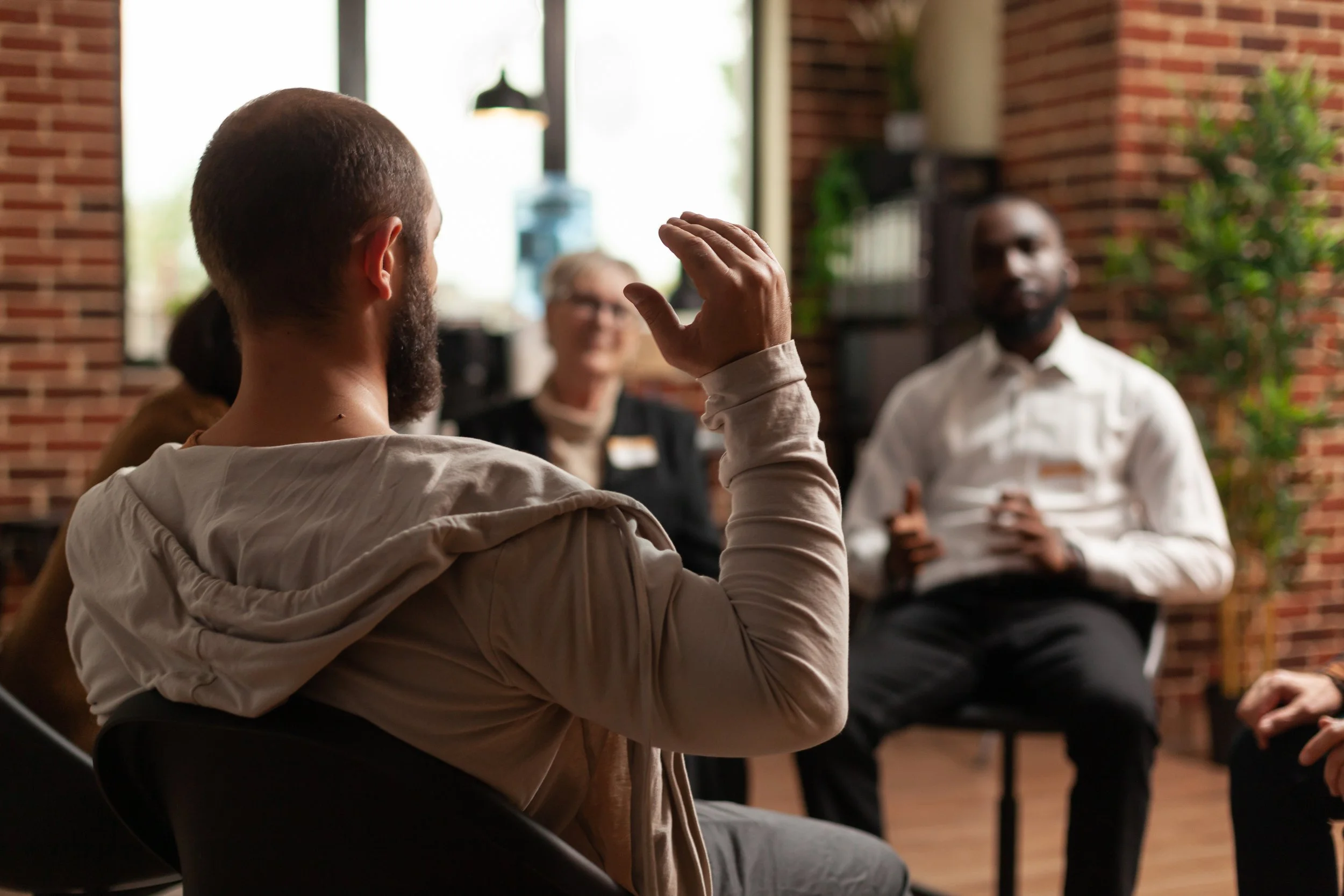 A man with a beard and short hair raising his hand during a discussion in a room with brick walls and large windows, people listening to him.