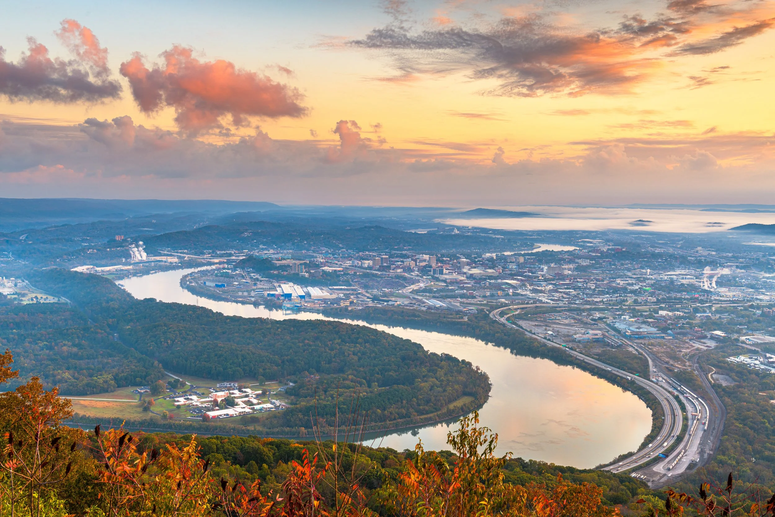 Aerial view of a city with a river, surrounded by hills and mountains, under a colorful sky with clouds during sunset or sunrise.