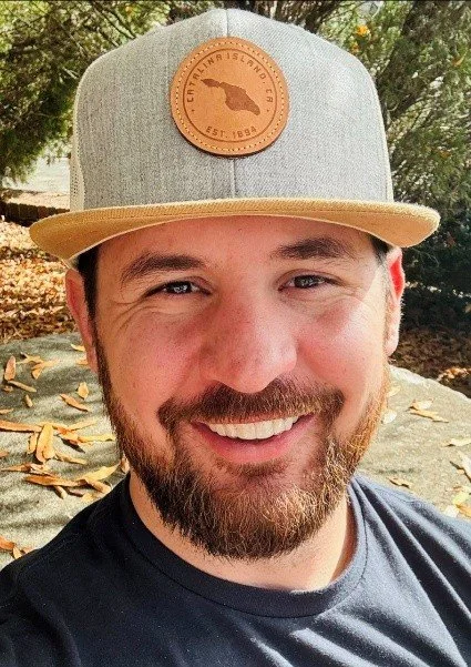 A smiling man with a beard wearing a gray baseball cap with a leather patch featuring a map of Florida and the words "City of Everglades, Florida, Est. 1984."