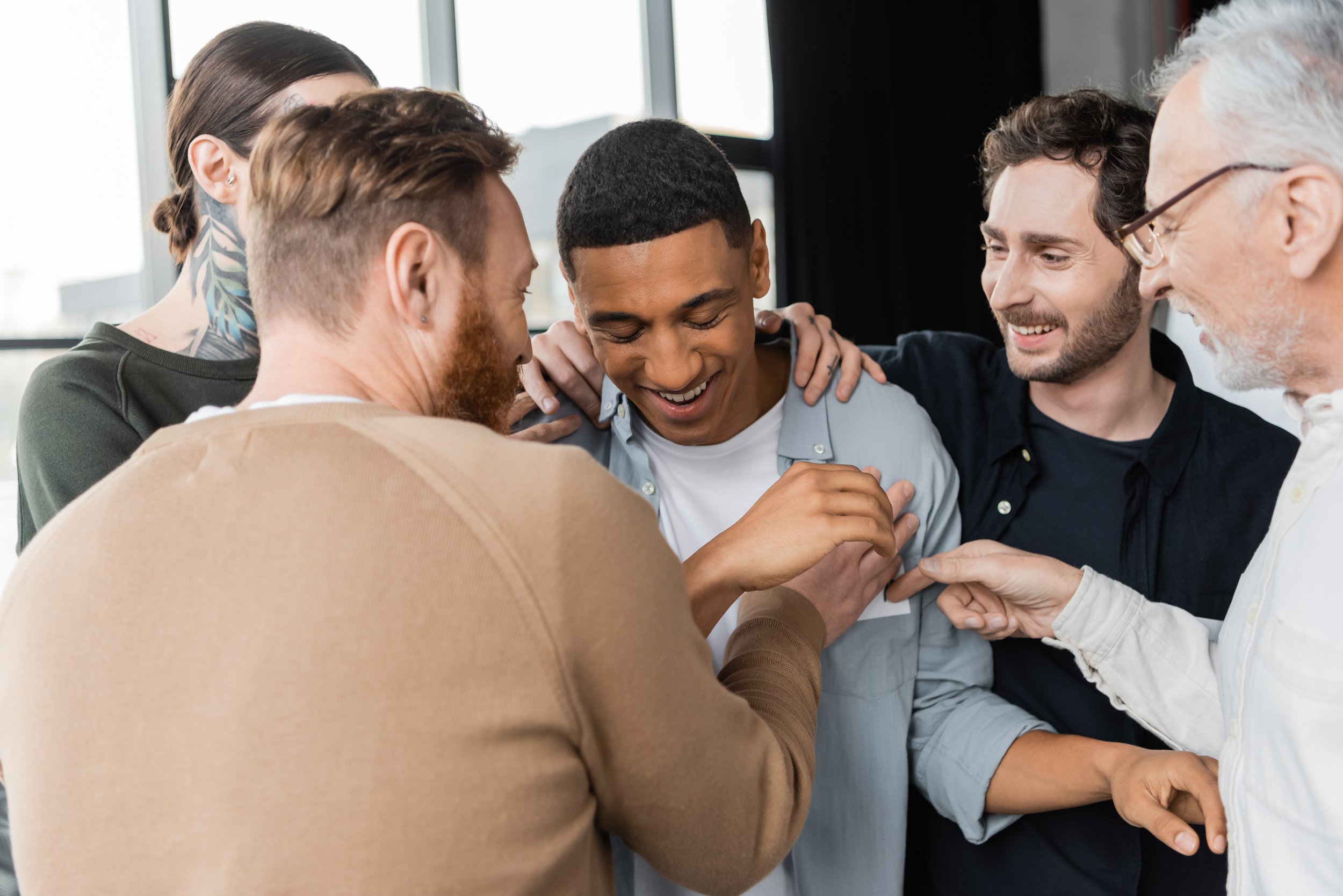 A group of five diverse people gathered closely, smiling, with their hands stacked together in a show of unity and teamwork in a bright room with large windows.
