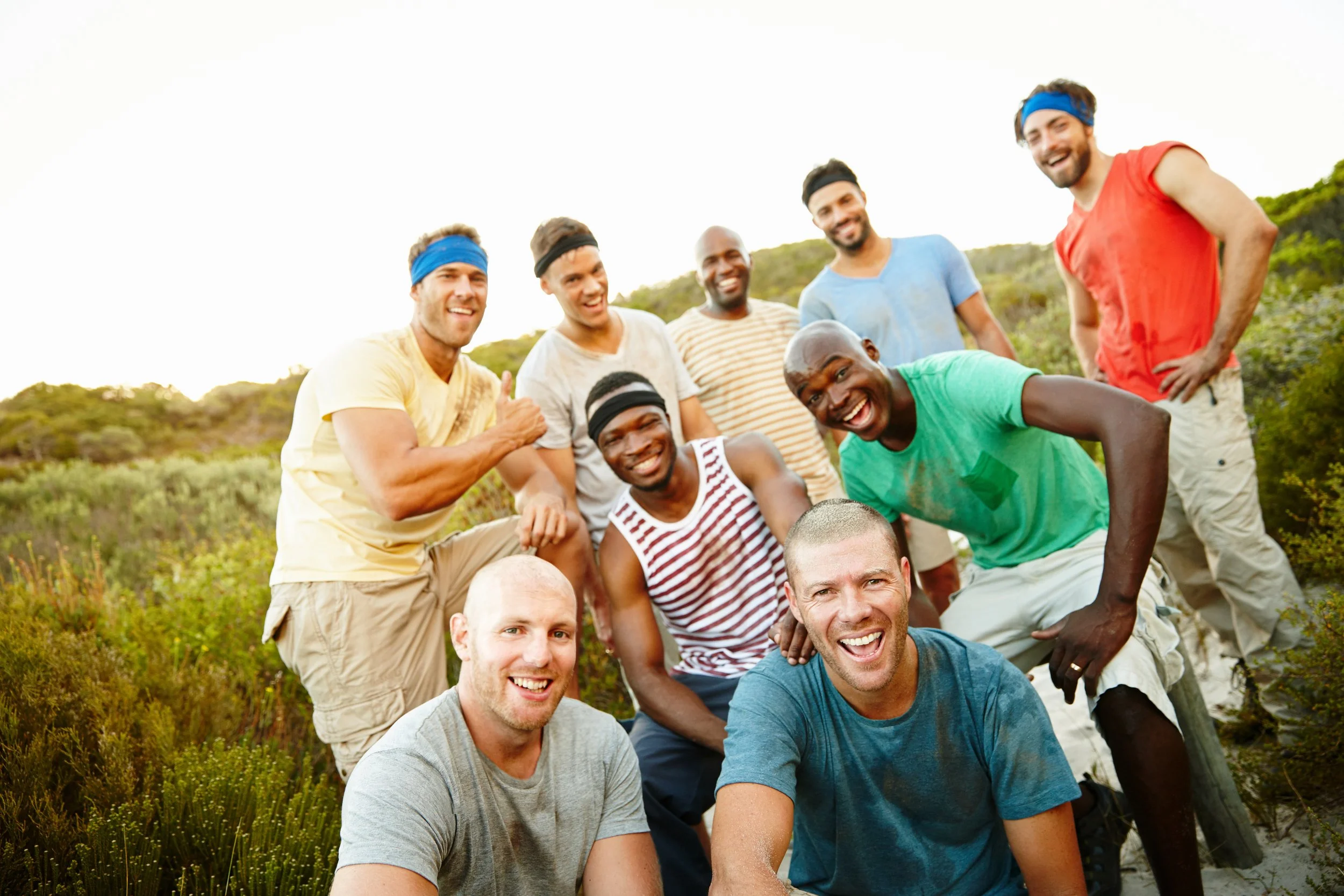 A group of smiling men, some wearing headbands, outdoors on a sunny day with greenery in the background.