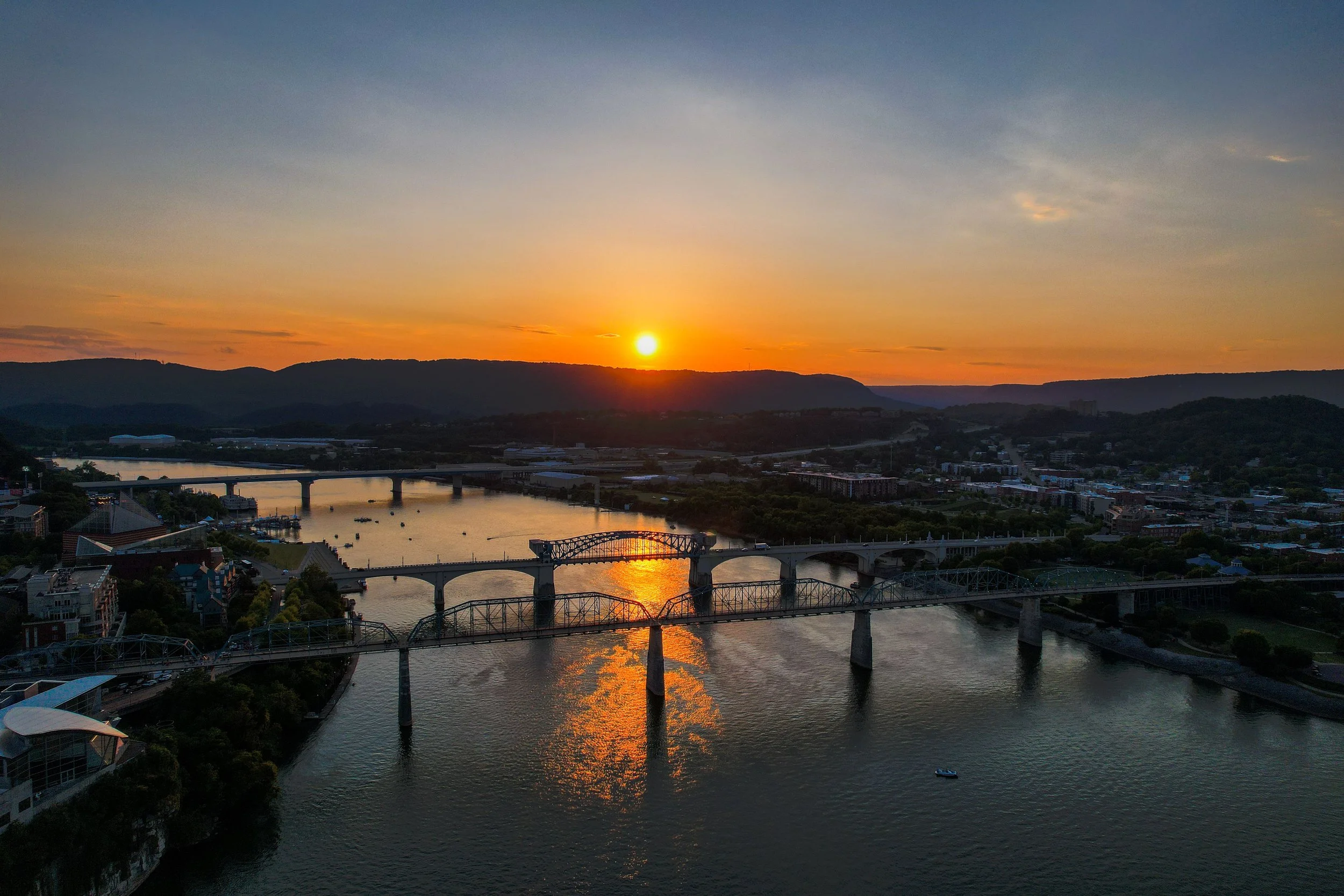 Sunset over a river with bridges, hills, and a cityscape in the background.