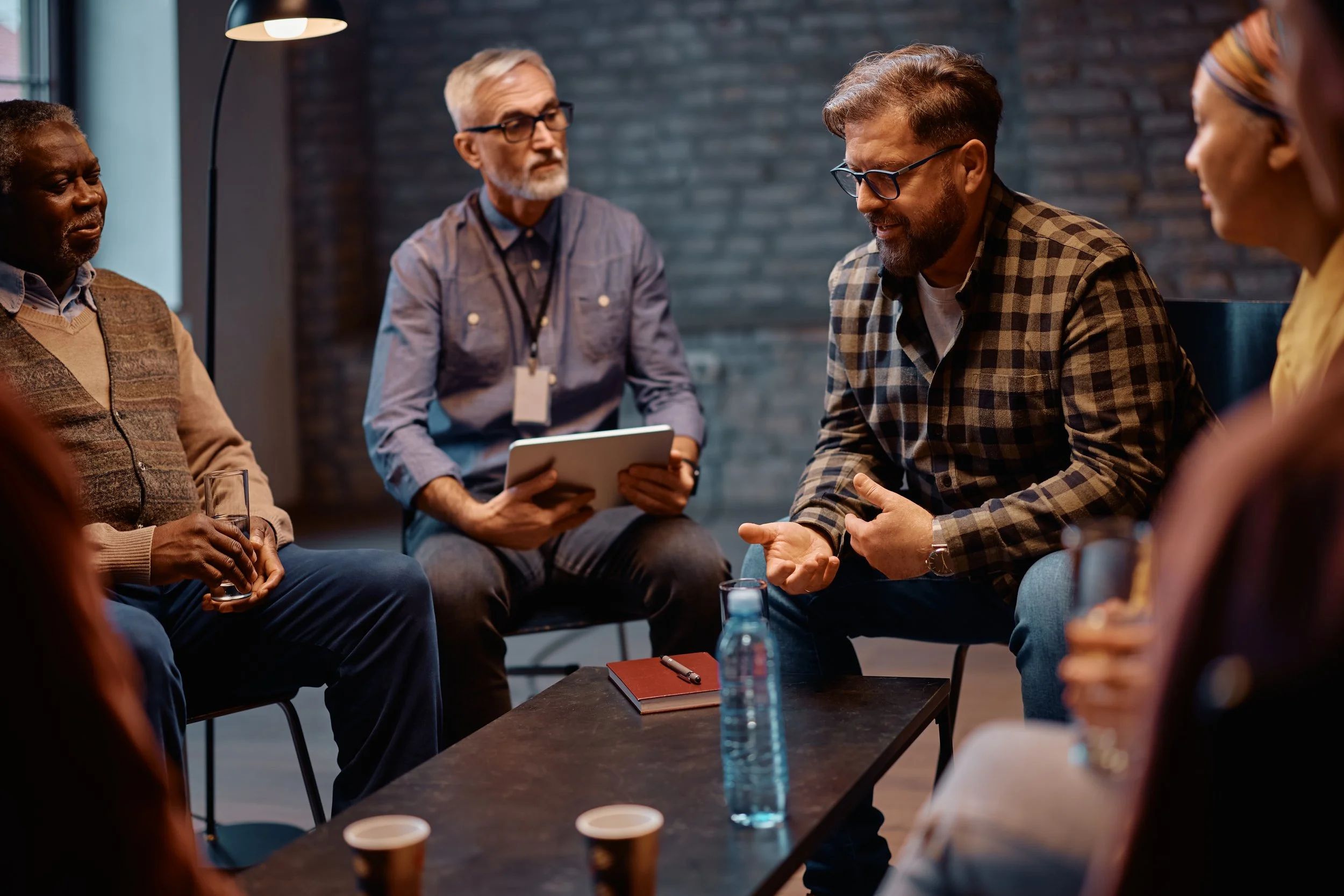 A group of diverse people gathered in a circular discussion, with a man in glasses and a plaid shirt speaking, others listening attentively, in a modern indoor setting with brick wall background.
