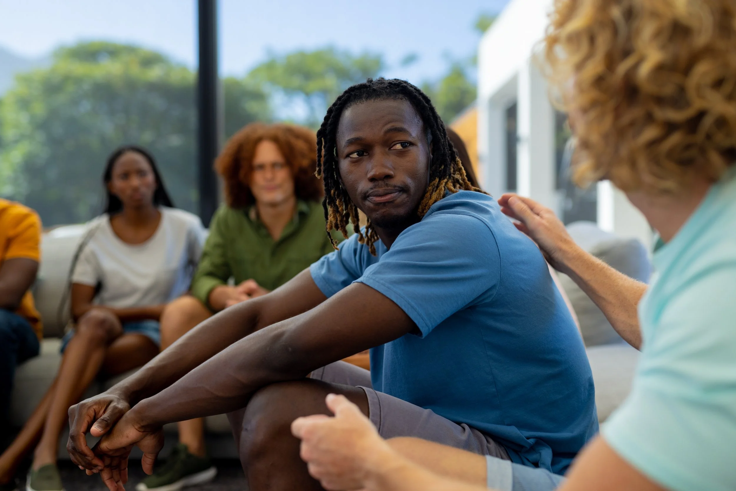 A group of diverse people sitting on the floor in a circle, engaging in conversation, with trees visible outside through large windows.